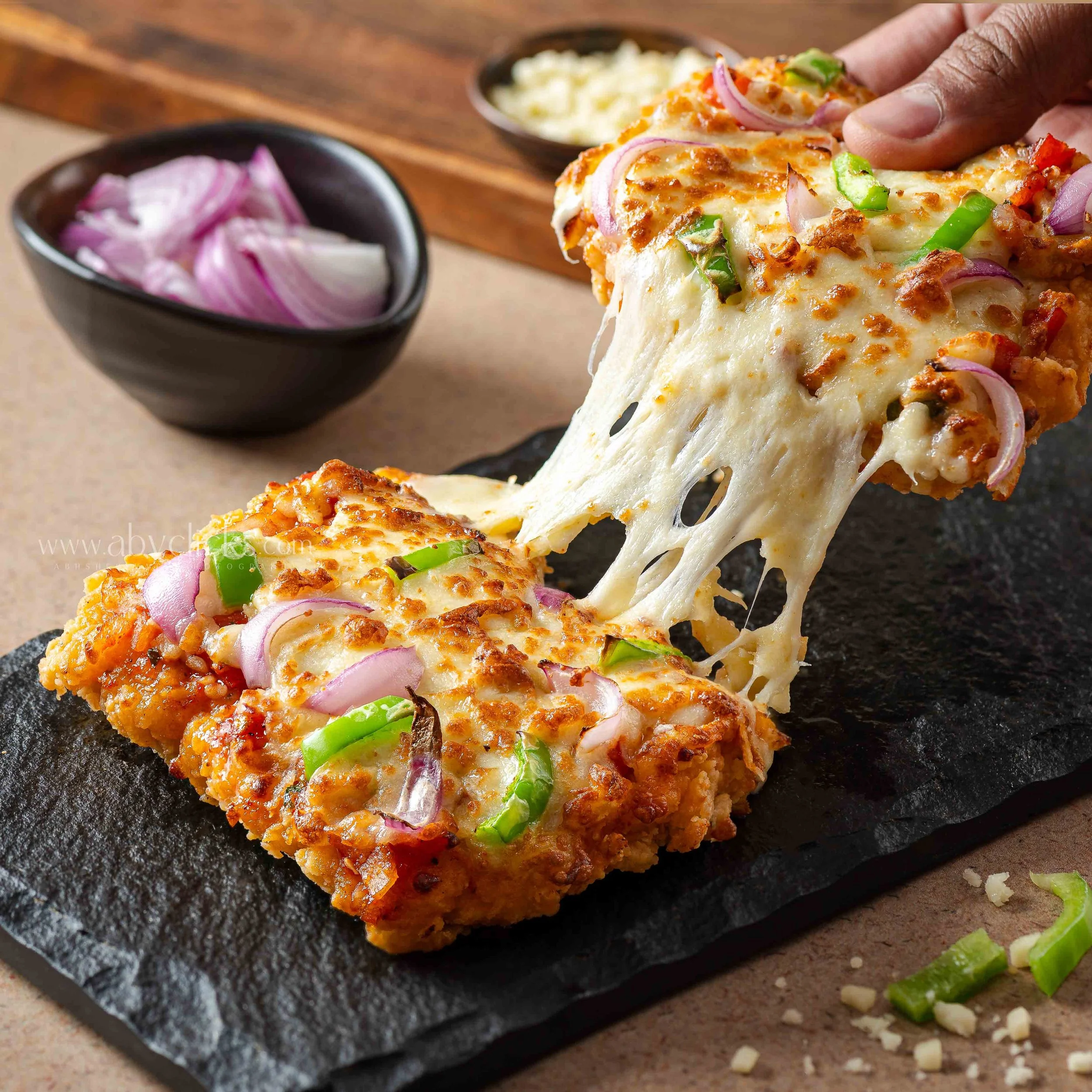 Close-up of a slice of cheesy pizza with green peppers and onions on a dark stone serving board.