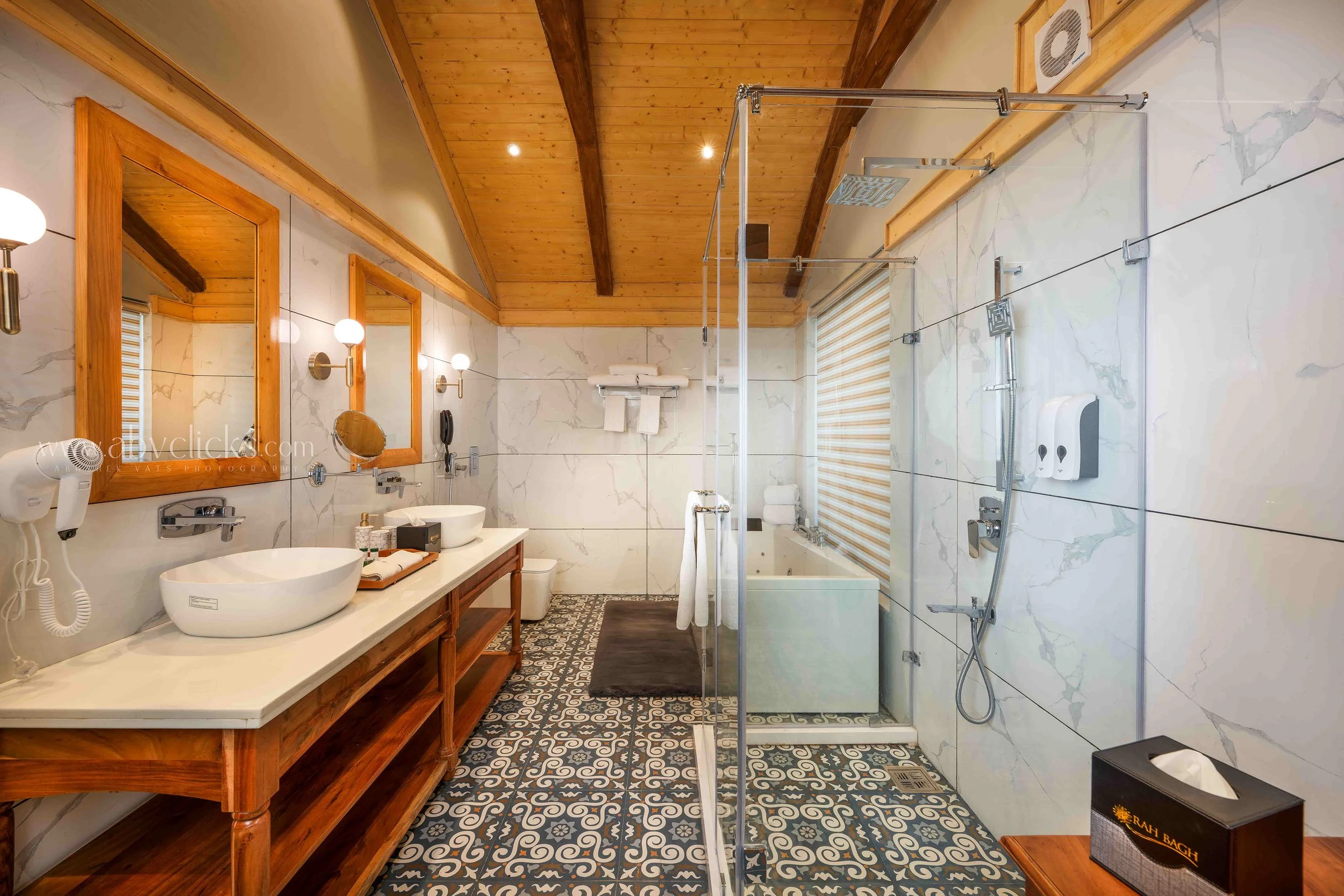 A rustic bathroom with a wooden ceiling and white marble tiled walls. It features a double sink vanity with wooden cabinetry, mirrors, wall-mounted lights, and a countertop with towels and toiletries. There is a walk-in shower with glass panels, a ra