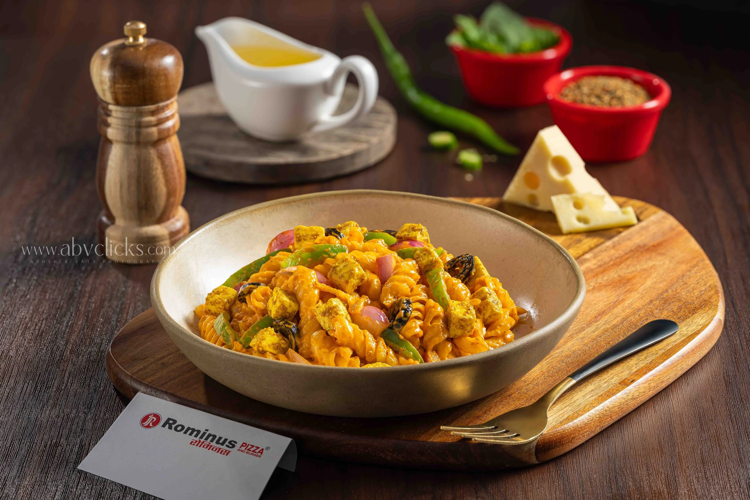 Bowl of pasta with vegetables and tofu on a wooden table, with cheese, green chili, and herbs in small bowls, salt shaker, and a business card in the background.
