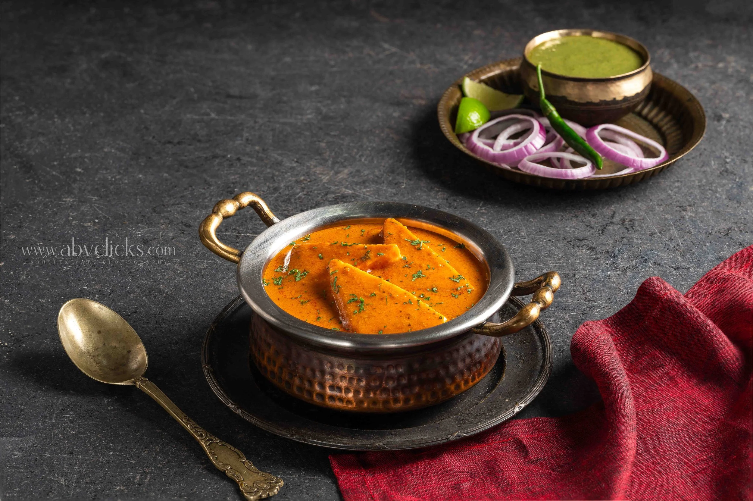 A bowl of Indian curry with pieces of bread, served in an ornate metal bowl on a black surface, with a decorative spoon nearby and a red napkin to the side. There is also a copper tray with sliced onions, lime, and green chili, and a small bowl of gr