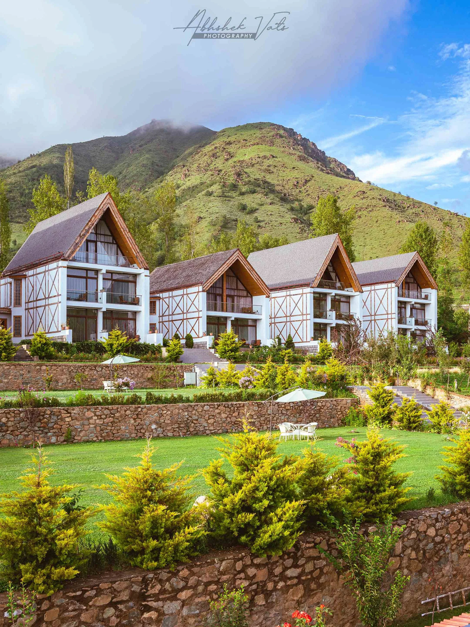A scenic view of a mountainous landscape with green hills and a blue sky with clouds. In the foreground, there are modern white buildings with wooden roofs, surrounded by well-maintained gardens, trees, and outdoor furniture.