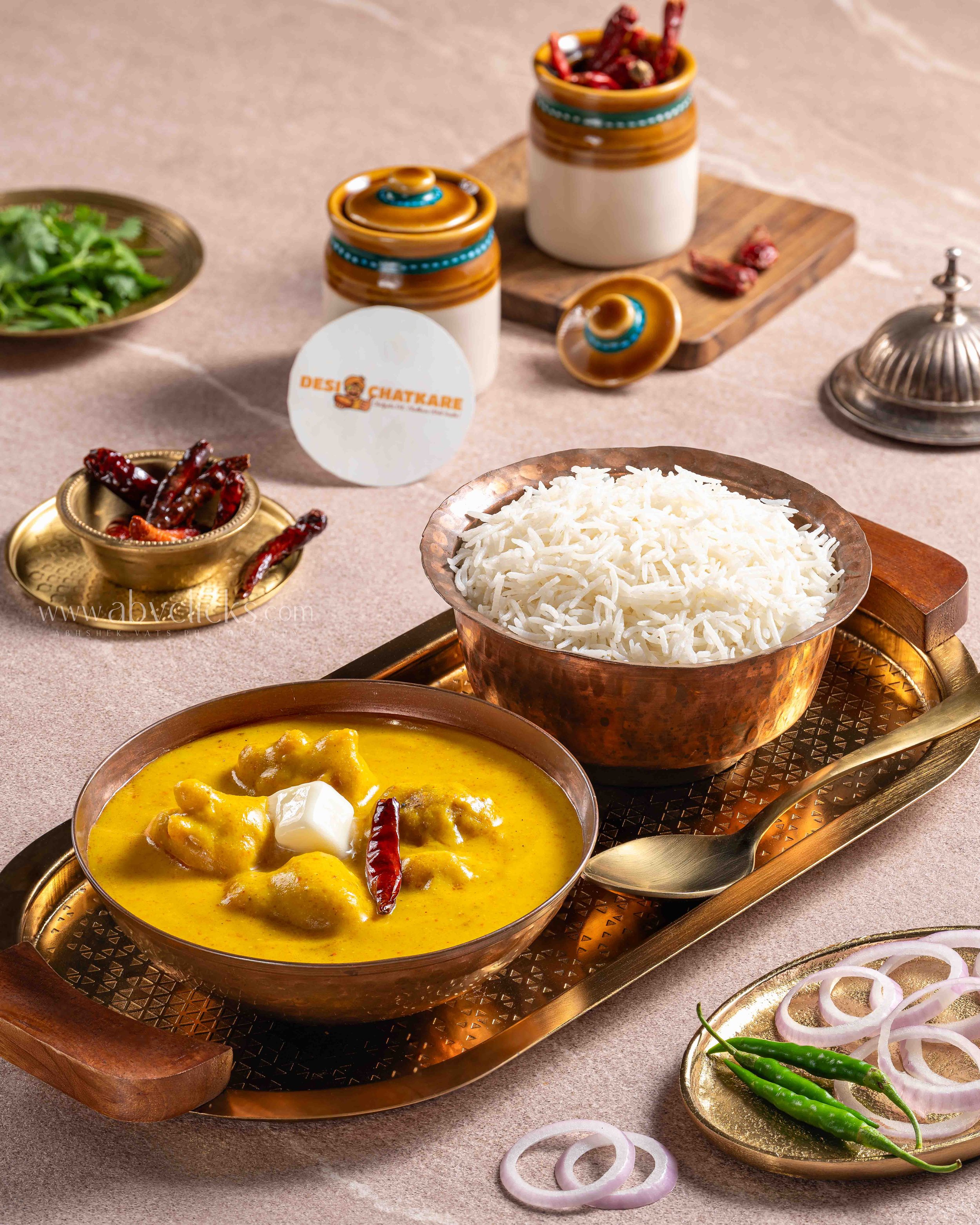 A traditional Indian meal with a yellow curry in a metal bowl, a bowl of white rice, a plate of sliced onions and green chilies, and containers of dried red chili peppers, all arranged on a copper tray with a wooden stand.