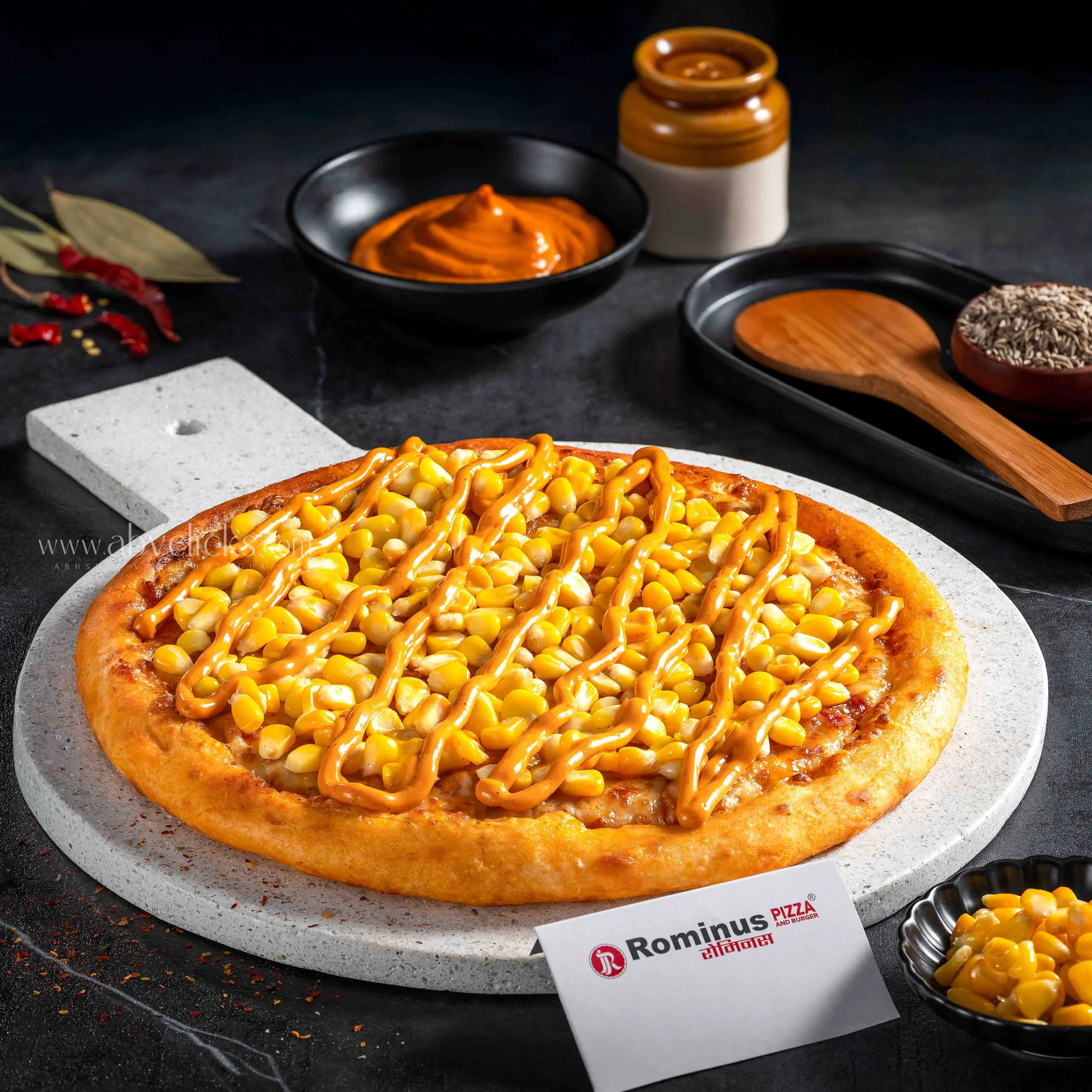 Close-up of a pizza topped with corn and drizzle of sauce on a white marble pizza board, surrounded by chili peppers and spice flakes, with a display tag and small bowl of corn beside it.