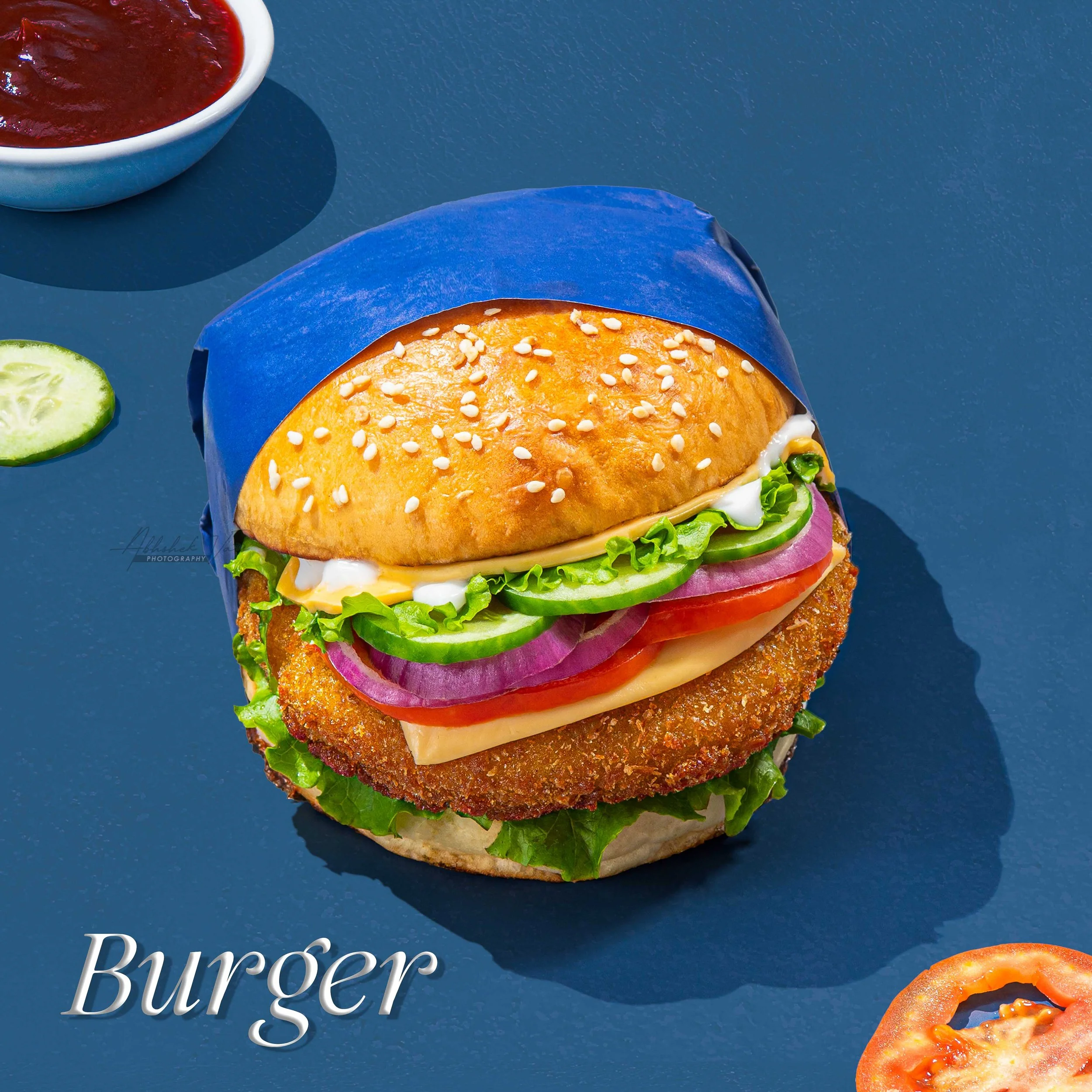 Close-up of a crispy chicken burger with lettuce, tomato, cucumber, onion, cheese, and sauce, wrapped in blue paper, on a blue surface with a bowl of ketchup and a sliced cucumber nearby.