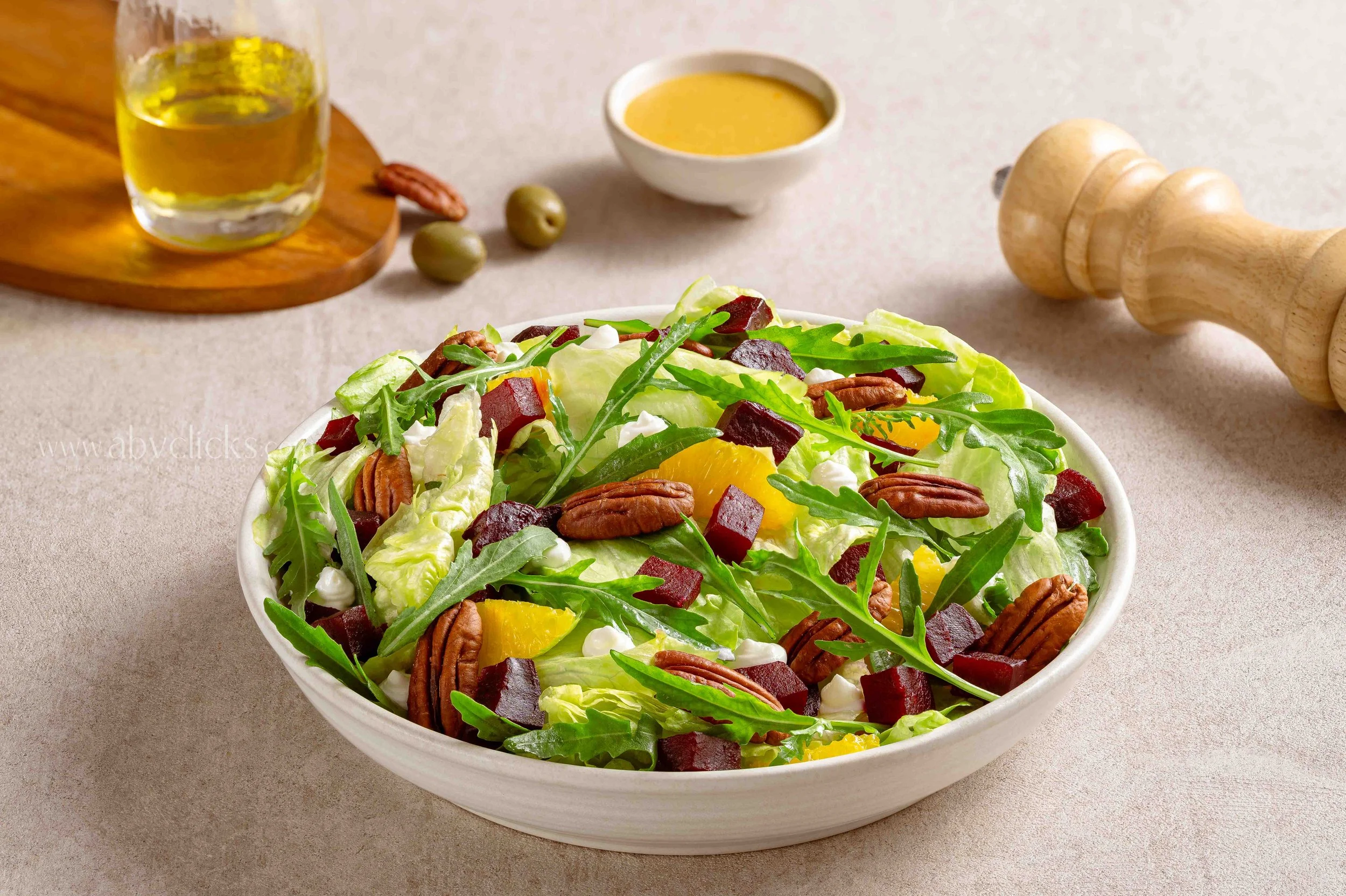 A bowl of fresh salad with lettuce, arugula, yellow bell peppers, beets, walnuts, and crumbled cheese, on a beige surface with a pepper grinder, bowl of dressing, and a glass of beer in the background.