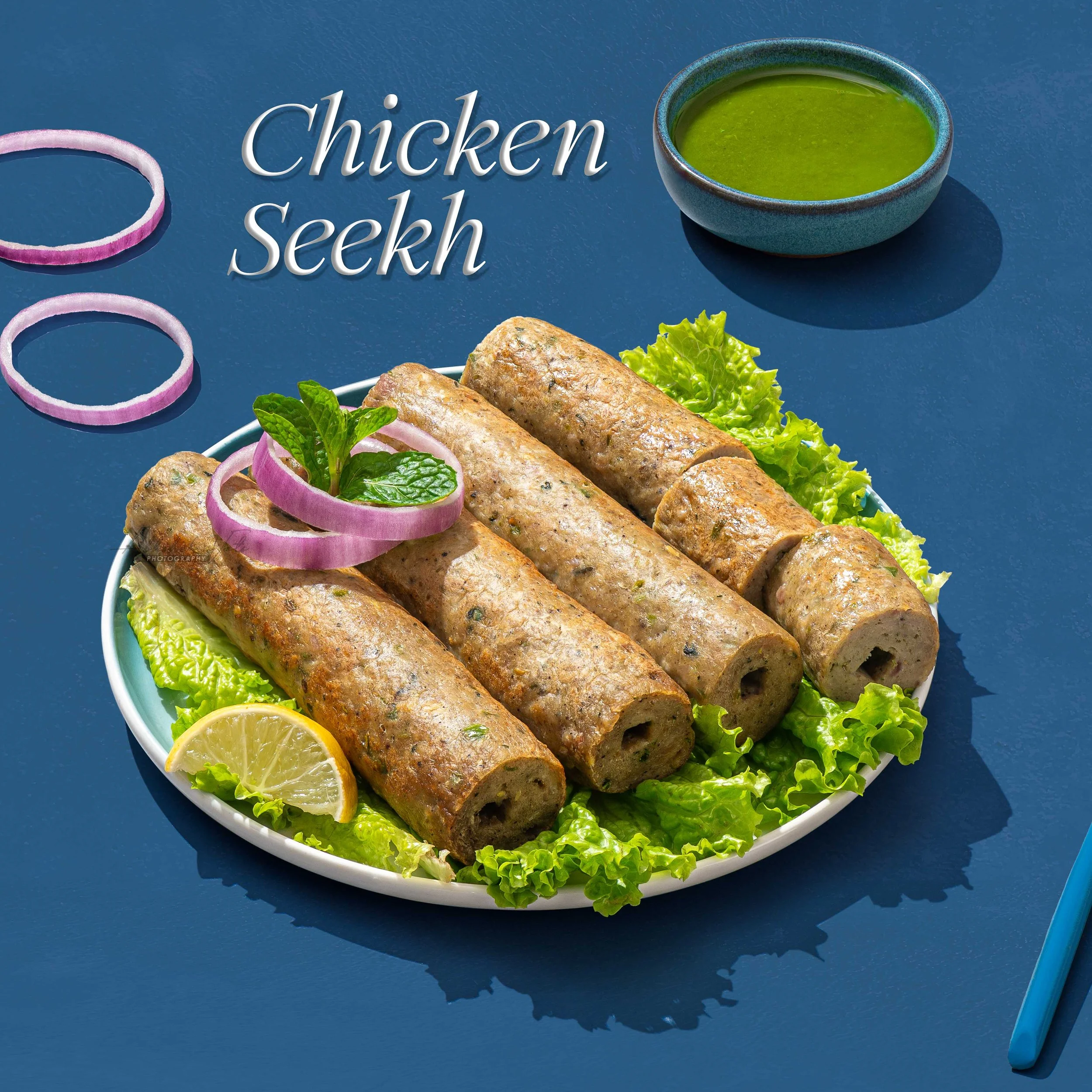 Plate of five cooked chicken sausages garnished with fresh green herbs and slices of lemon, with a side of green dipping sauce in a bowl and some onion rings and a spoon on a blue surface.