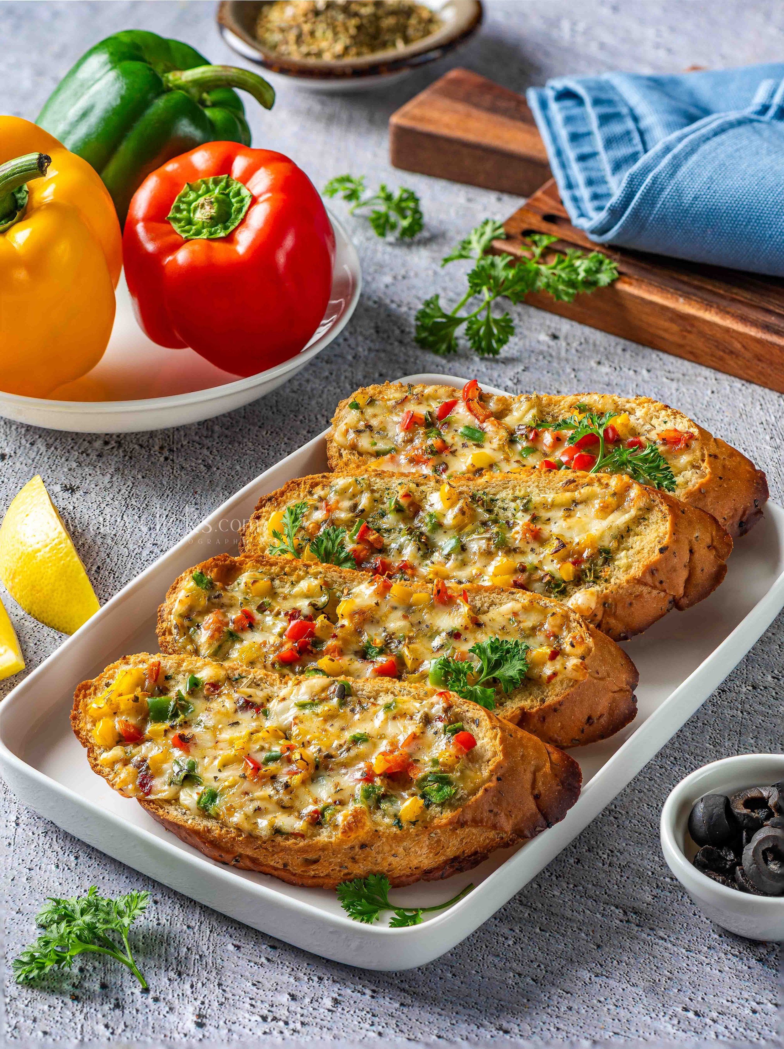 Three slices of cheesy garlic bread topped with chopped red and green bell peppers and herbs on a white rectangular plate, with fresh parsley garnished and a lemon wedge, on a textured gray surface.