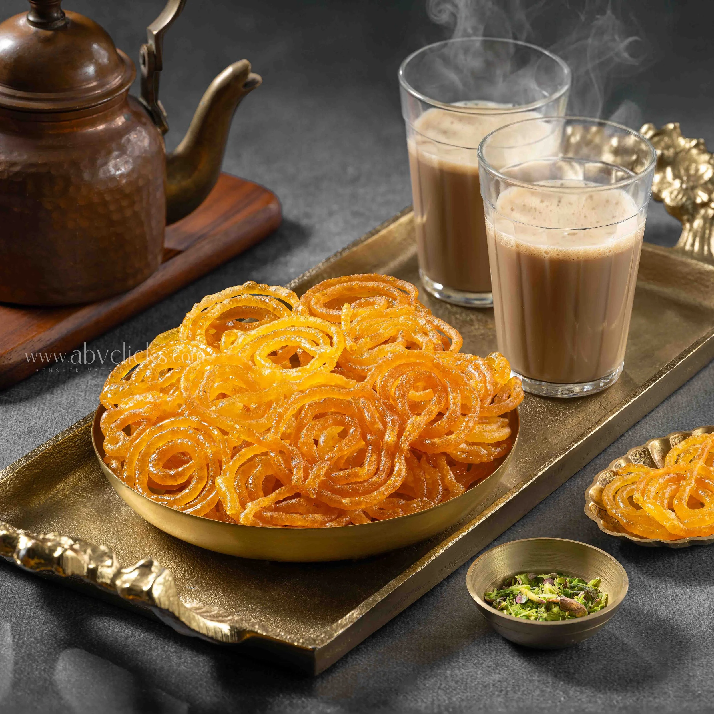 A tray with two glasses of chai, a copper kettle, and a bowl of chopped green pistachios on a dark table.