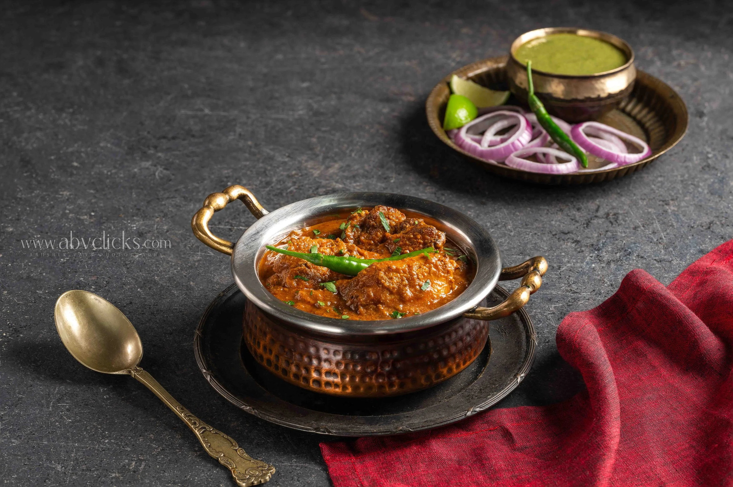 Traditional Indian curry served in copper bowl garnished with green herbs, with onion rings, lime wedges, and green chili on a decorated tray, accompanied by a gold spoon and red cloth napkin on a dark surface.
