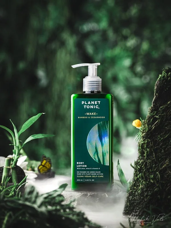 Green bottle of Planet Tonic Wake body lotion with bamboo and cedarwood scent, positioned among lush greenery and rocks, with a foggy or misty background.