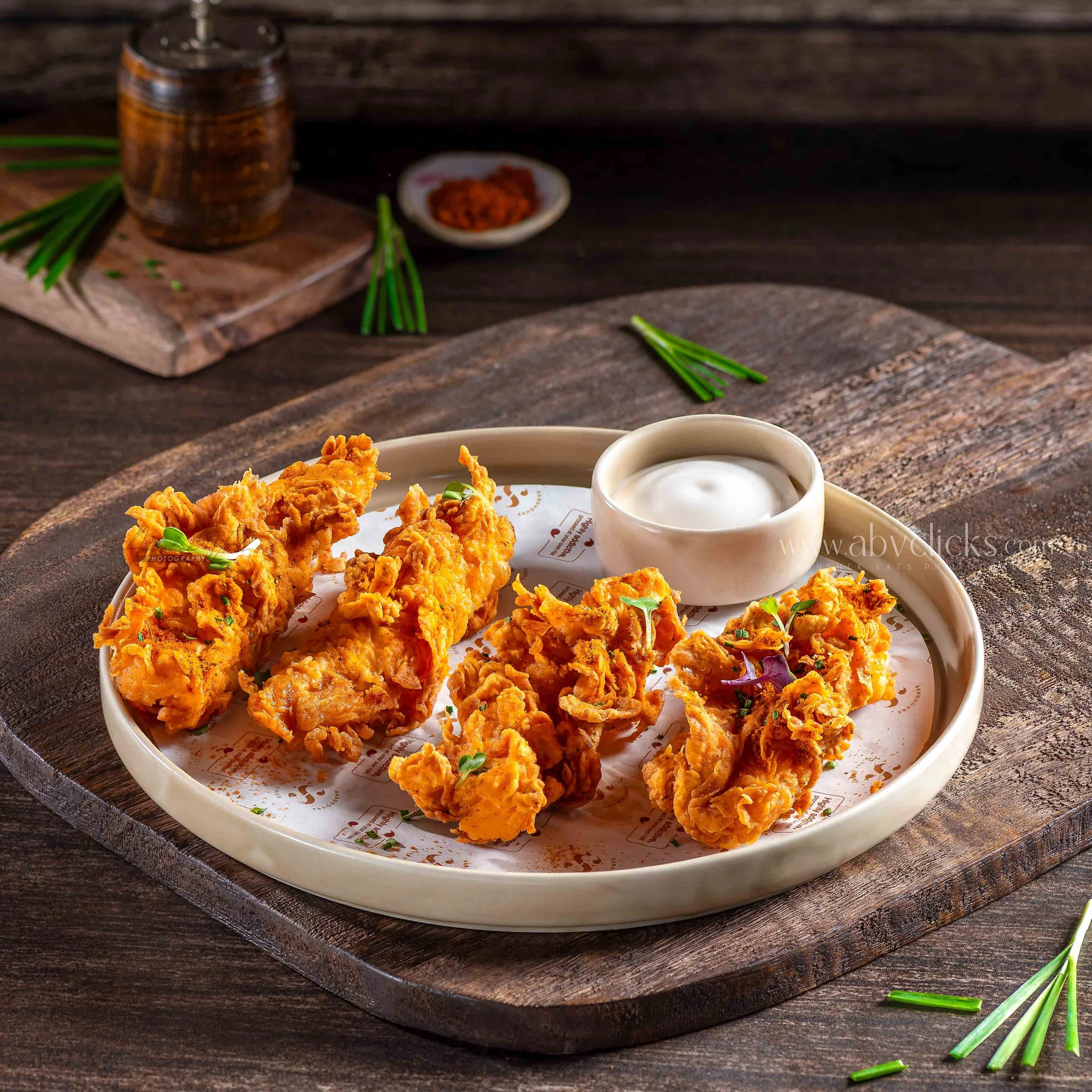 Plate of crispy fried chicken with a cup of dipping sauce on a rustic wooden table, with a small bowl of chili flakes and green garnish nearby.