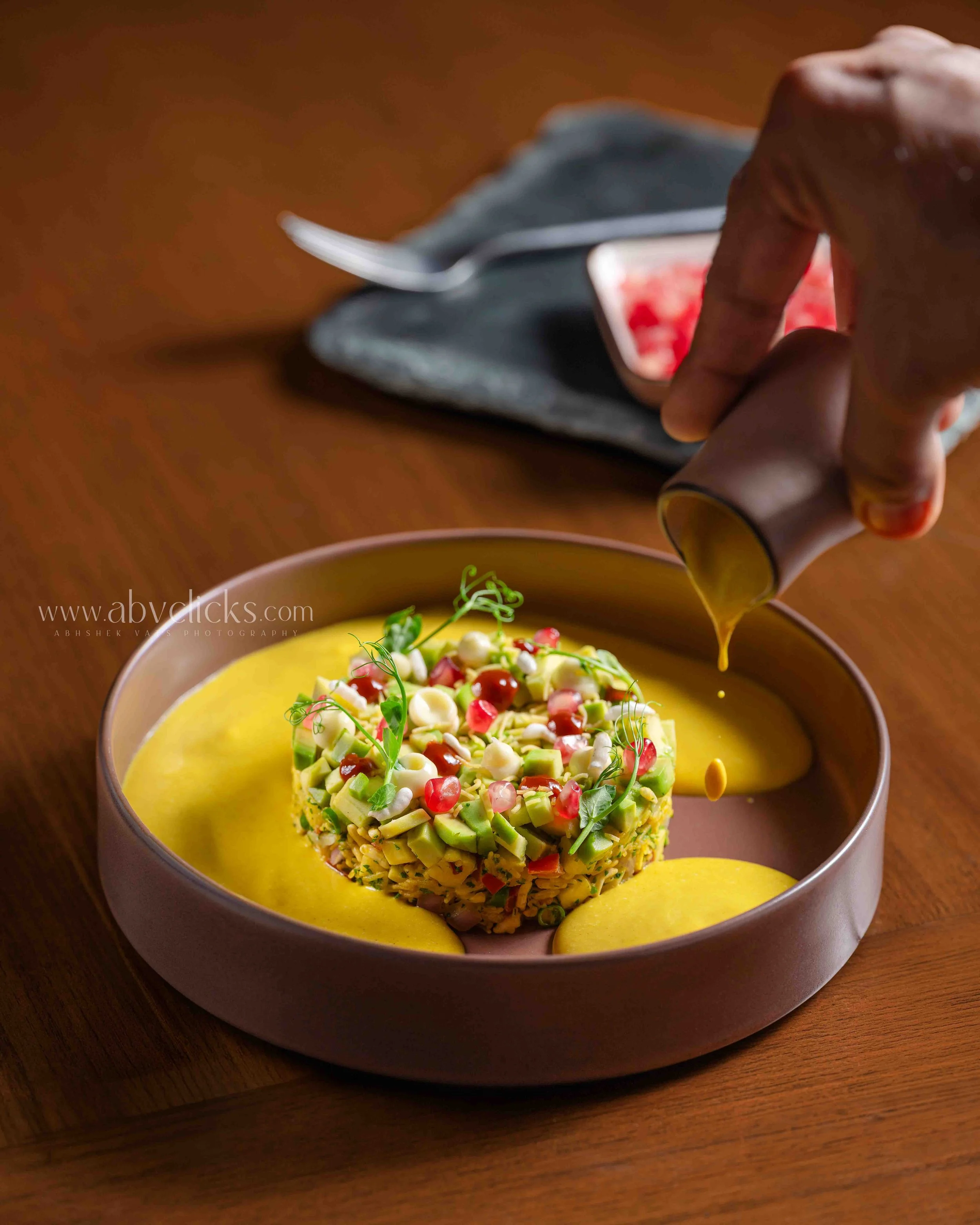 A bowl with a yellow liquid dish and a dessert topped with chopped nuts, pomegranate seeds, and microgreens on a wooden table.