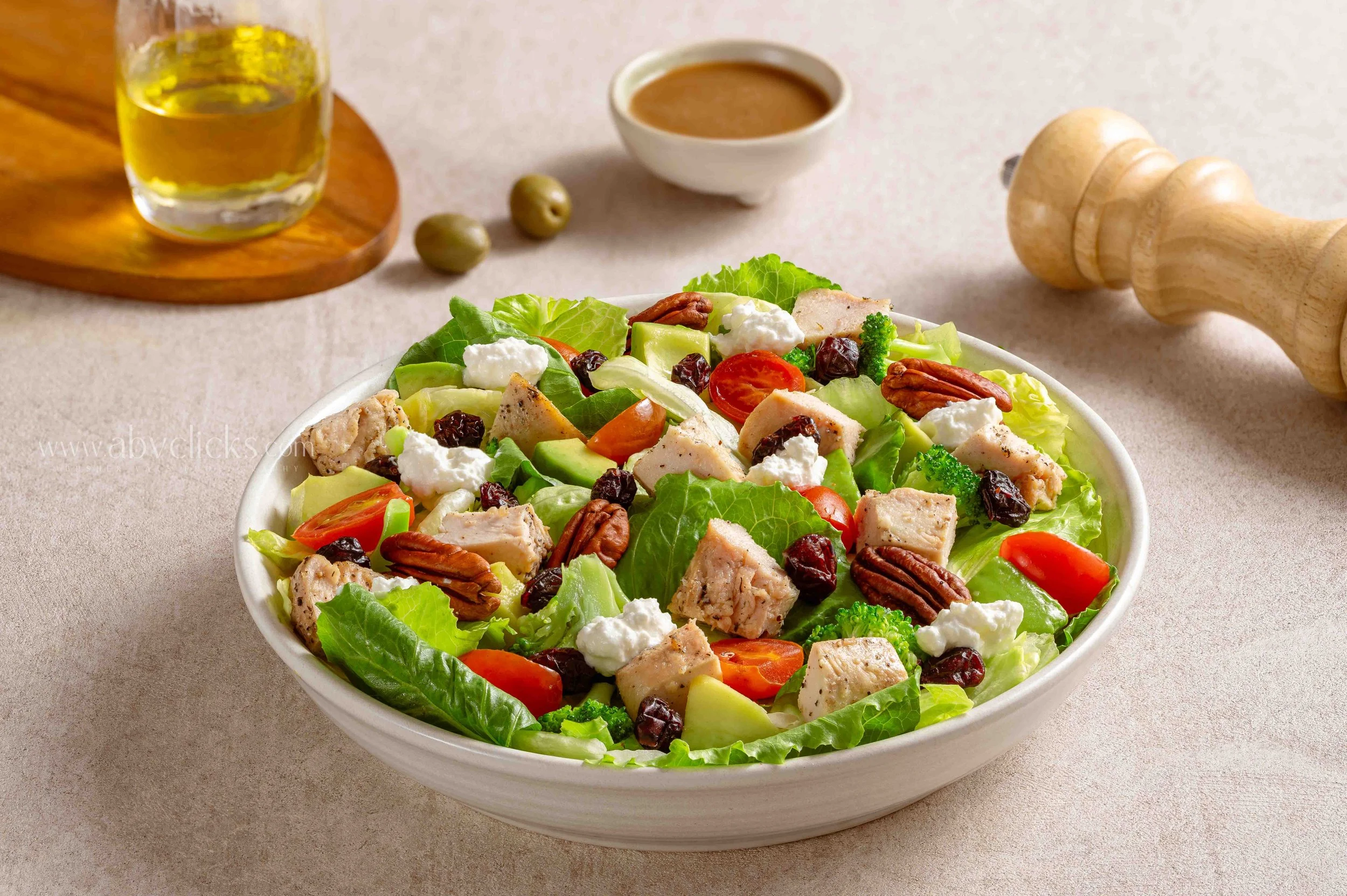 A large white bowl filled with a vegetable salad containing lettuce, cherry tomatoes, cucumbers, pecans, dried cranberries, grilled chicken pieces, and dollops of feta cheese, on a light-colored table.