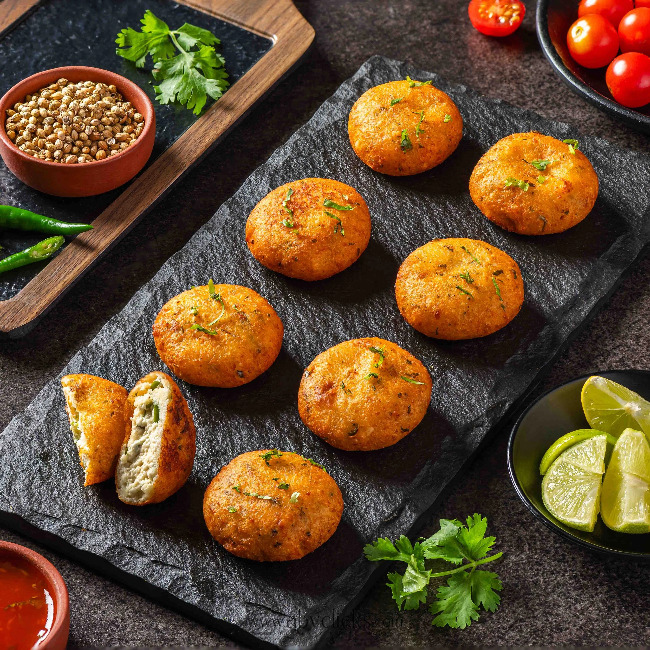 Fried veggie balls garnished with cilantro on a black slate serving platter, with fresh tomatoes, lime wedges, and green chilies nearby, on a dark countertop.