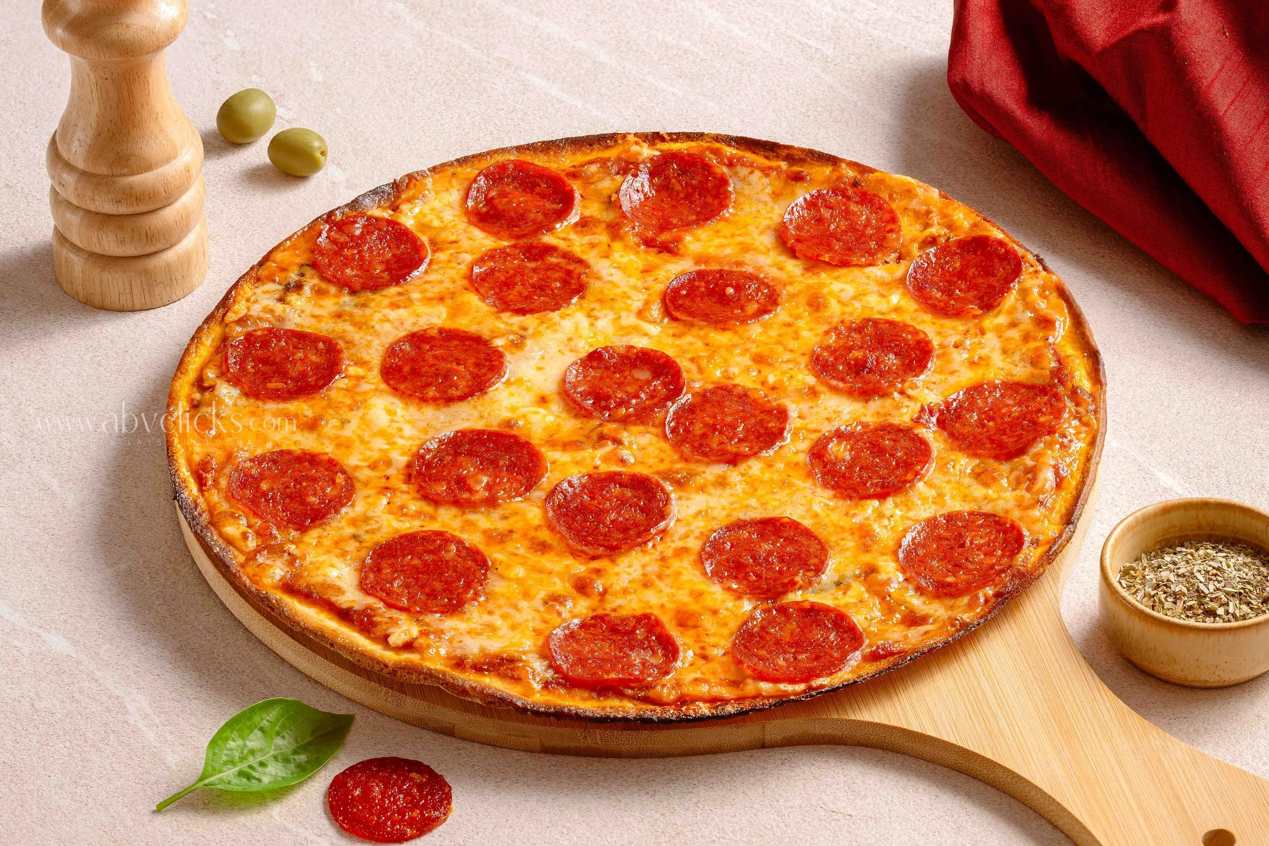 A pepperoni pizza on a wooden pizza board with ingredients and utensils around it.