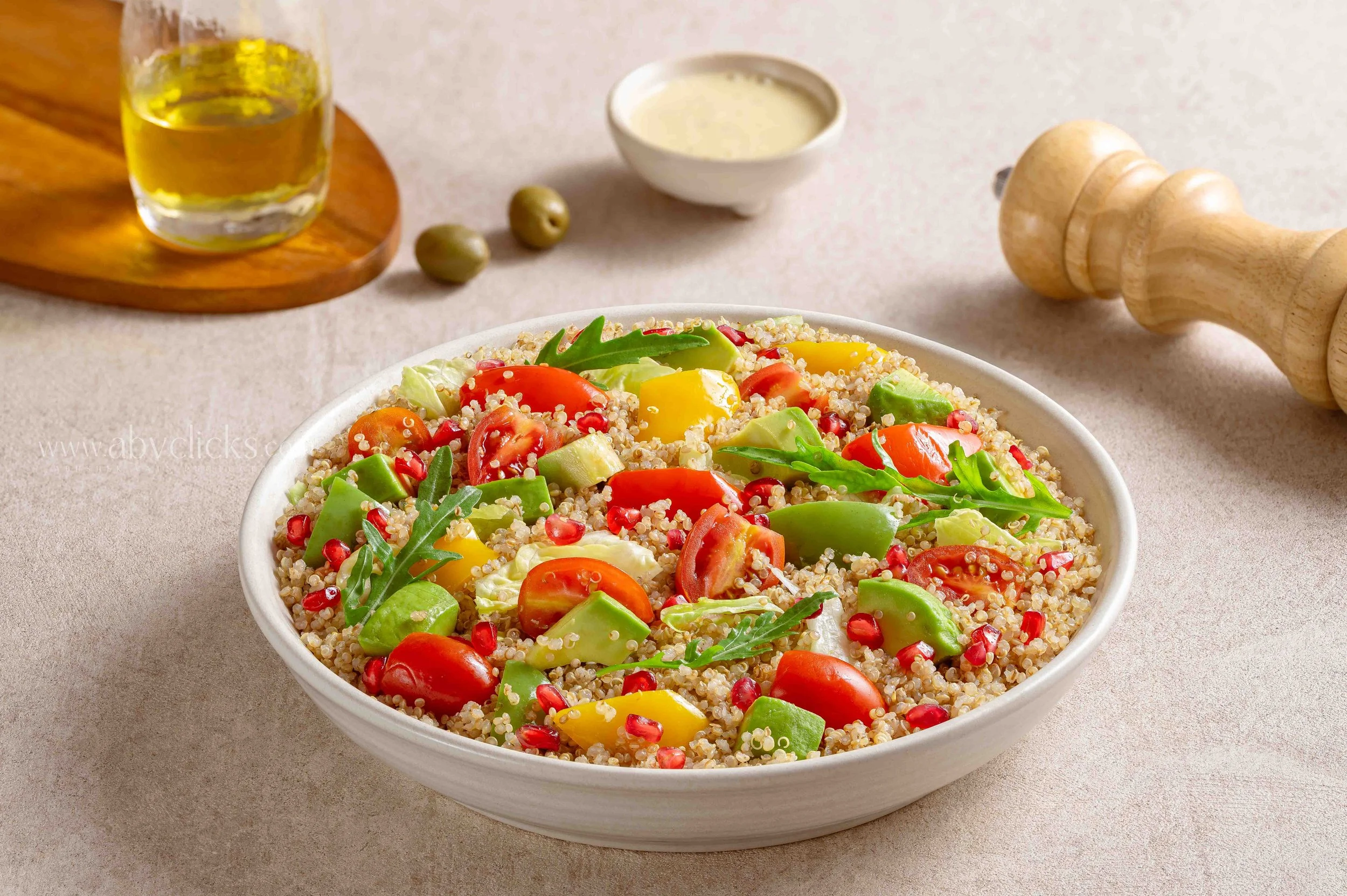 A bowl of quinoa salad with cherry tomatoes, avocado, and arugula, on a light-colored surface, with a wooden pepper mill, a small bowl of dressing, a glass of olive oil, and green olives in the background.