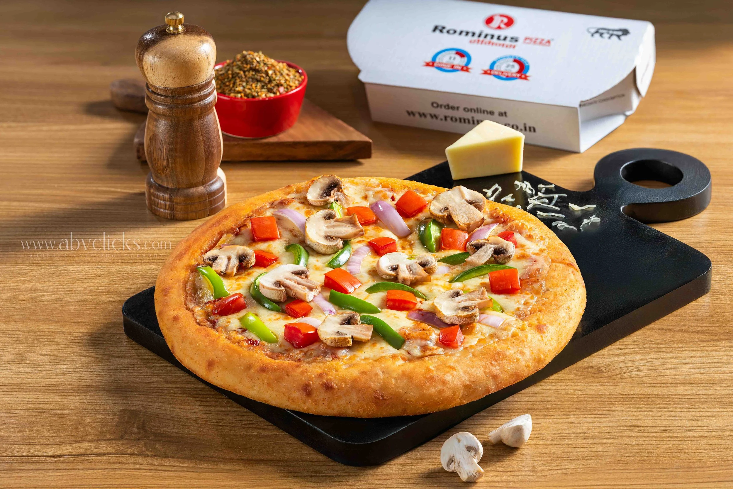 A freshly baked pizza topped with sliced mushrooms, red and green bell peppers, and red onions on a black pizza tray, placed on a wooden table. In the background, there is a package of Romiwas pizza, a wedge of cheese, a black pizza cutter, a wooden 