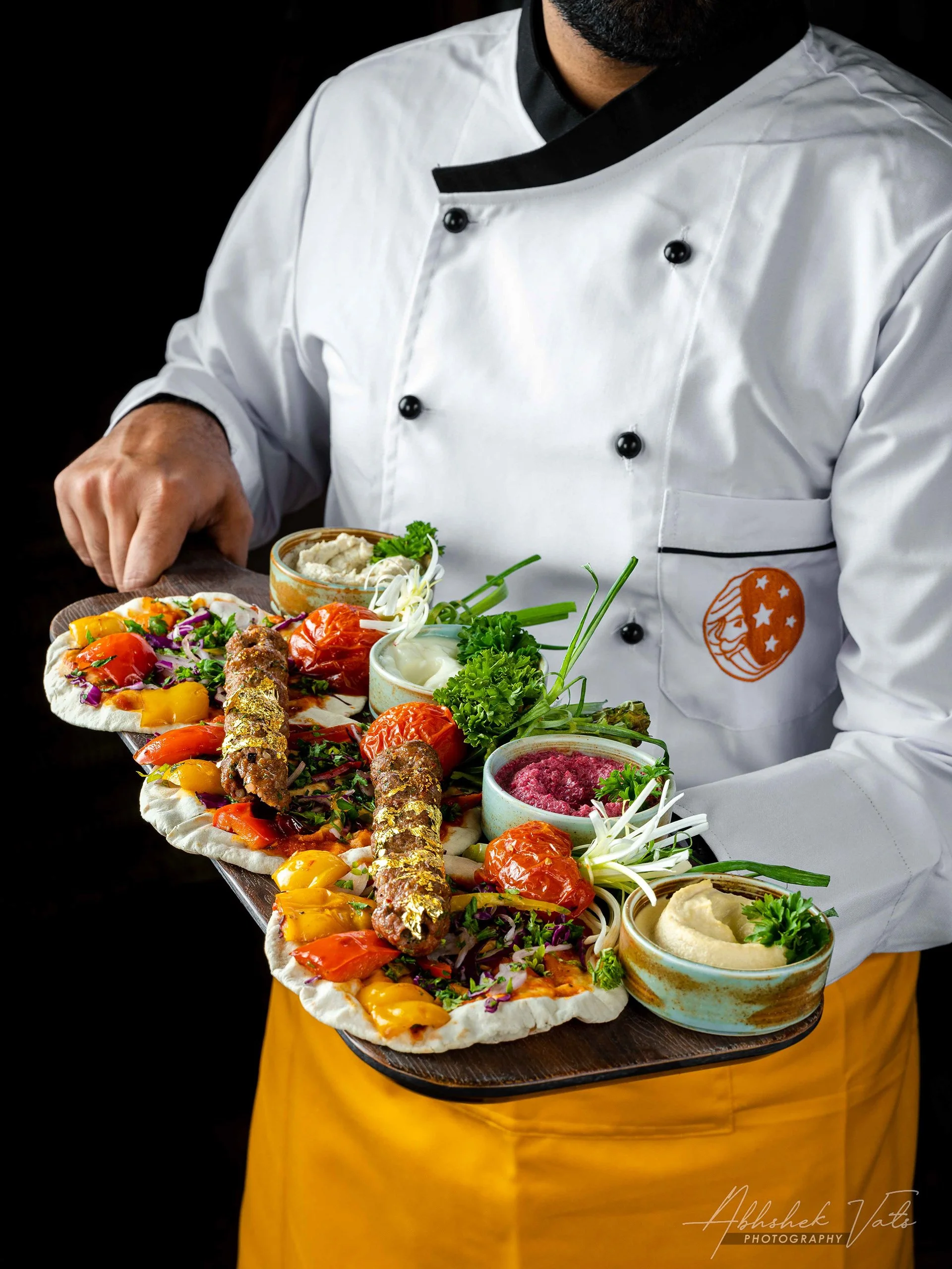 A chef in a white and black chef's coat serving a variety of colorful dishes on a wooden tray, including vegetables, sauces, and garnishes, against a dark background.