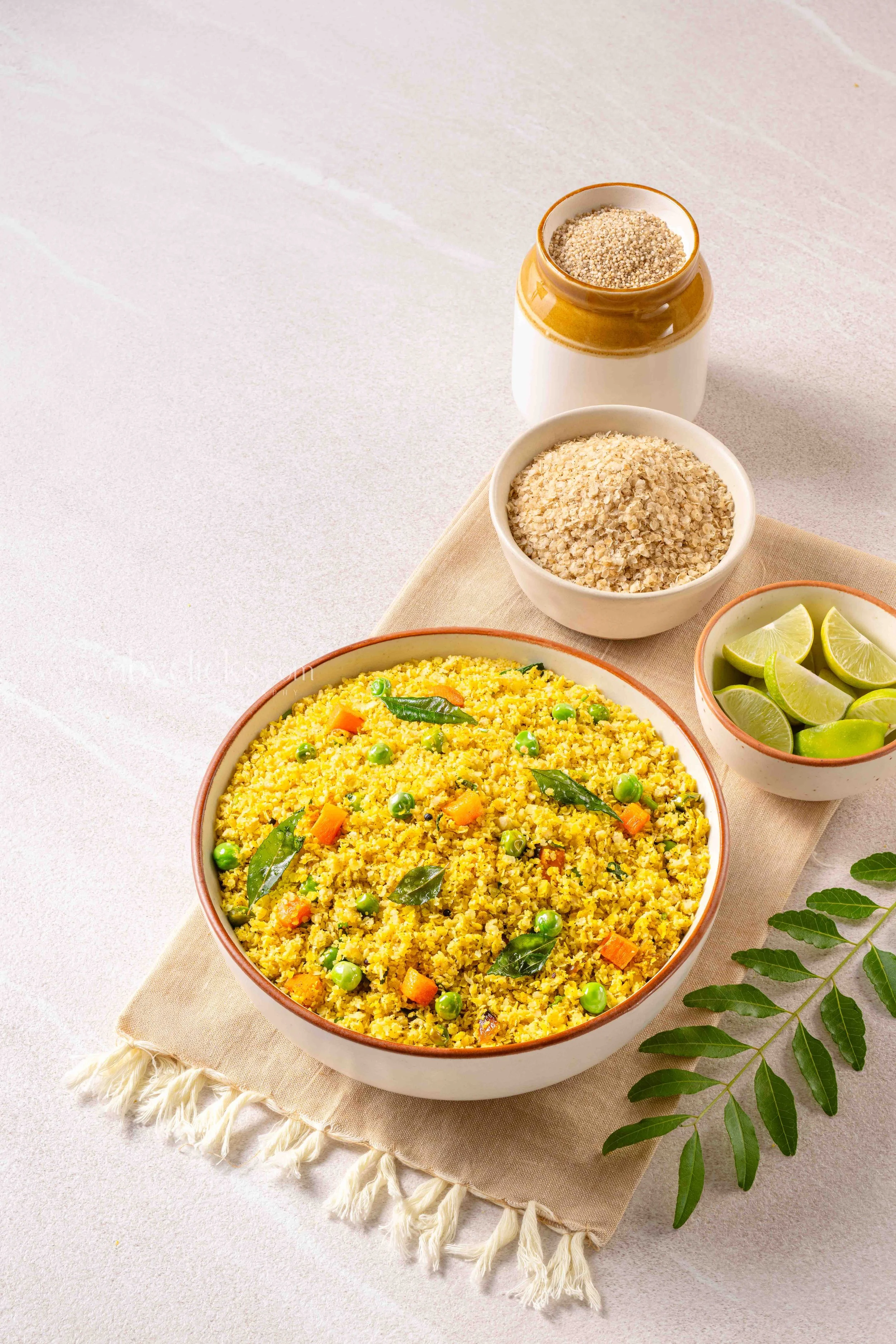Bowl of cooked yellow Indian-style rice with peas and carrots, garnished with fresh curry leaves, set on a beige cloth napkin with lime wedges and green leaves nearby.