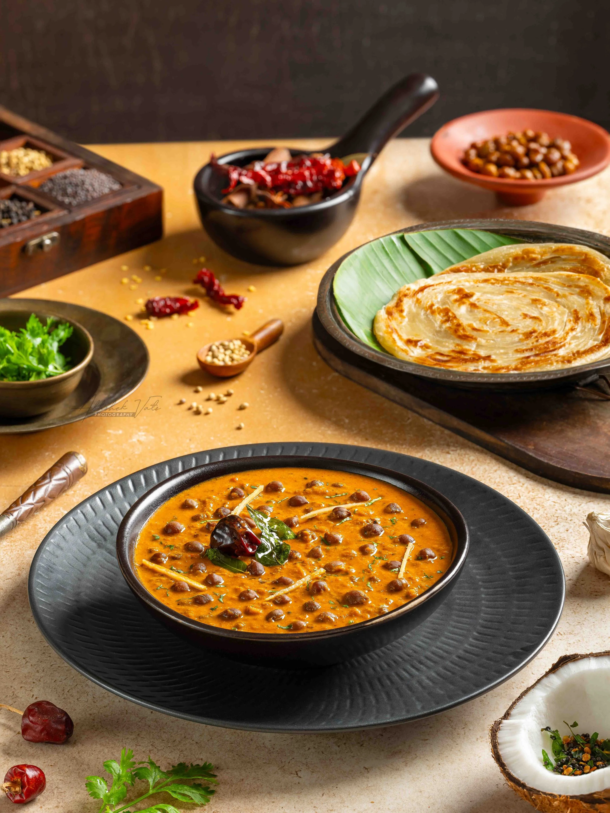 A bowl of Indian lentil soup garnished with herbs and spices, surrounded by various dishes, spices, and ingredients on a beige surface.