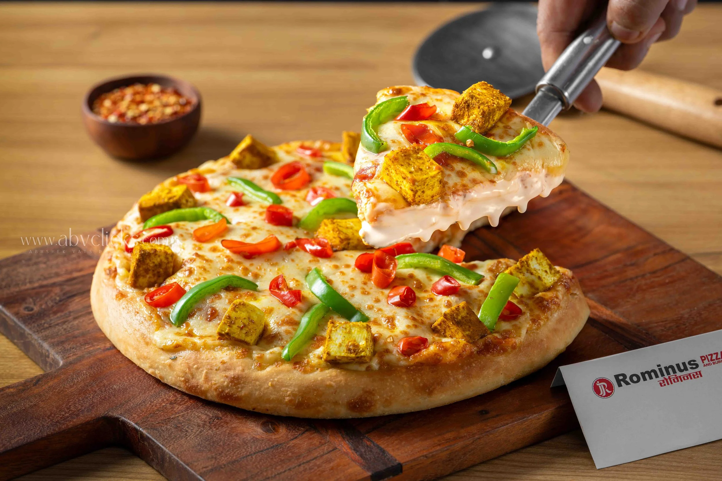 A freshly baked pizza with cheese, green bell peppers, red peppers, and pieces of seasoned paneer being placed on a wooden surface by a pizza cutter. In the background, there is a small bowl of crushed red pepper flakes and a partially visible pizza 