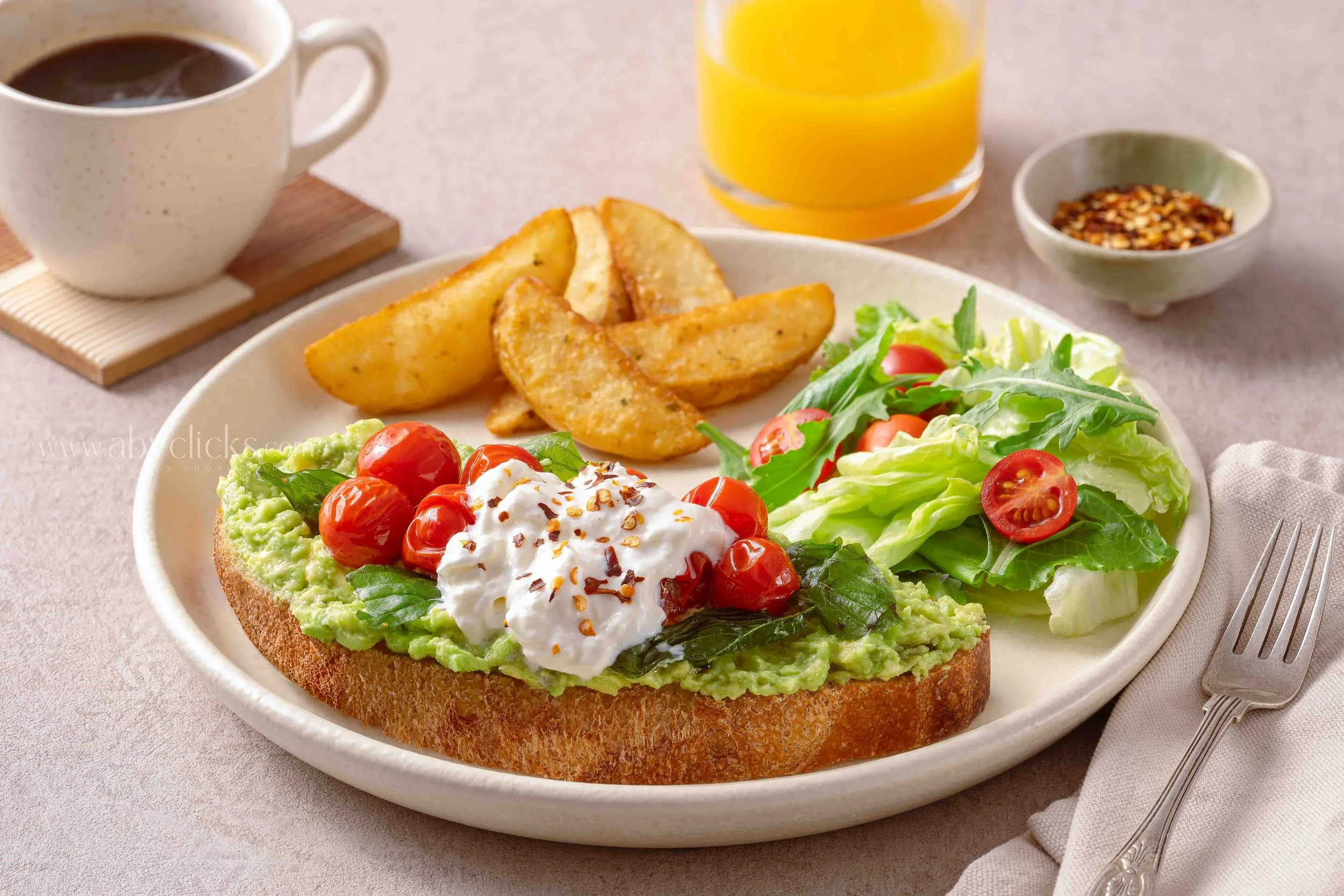 A plate with toasted bread topped with mashed avocado, cherry tomatoes, a dollop of sour cream, and sprinkled red pepper flakes, accompanied by a side salad of lettuce, cherry tomatoes, and greens, with a glass of orange juice, a cup of coffee, and a small bowl of chili flakes in the background.