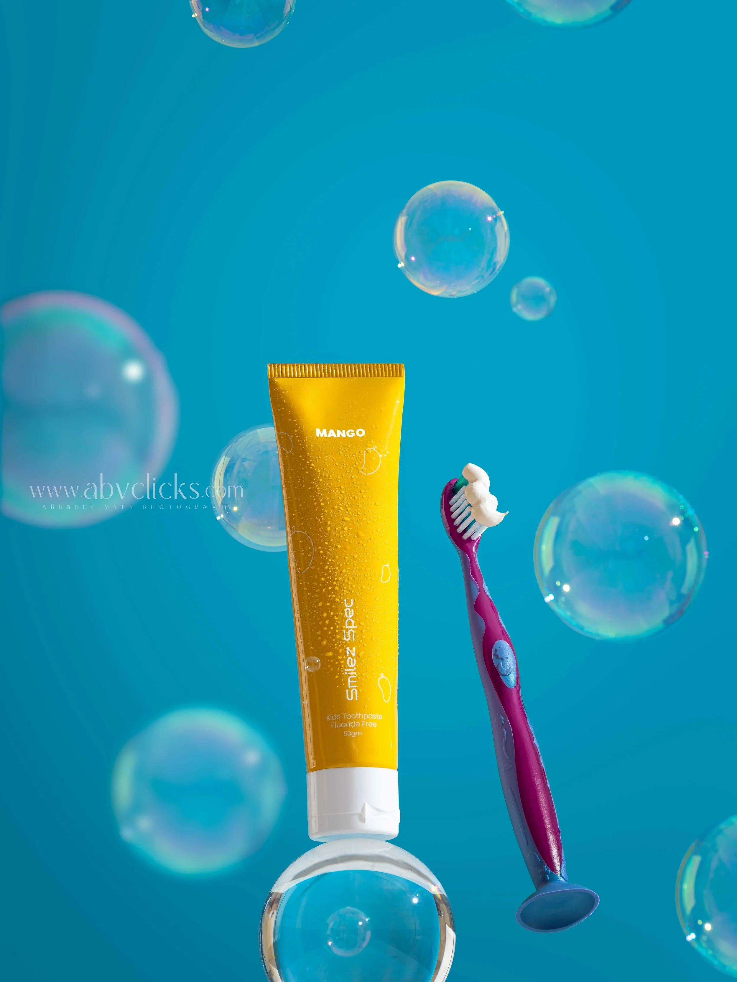 A yellow kids' toothpaste tube labeled 'MANGO' with a toothbrush, surrounded by soap bubbles and a blue background.