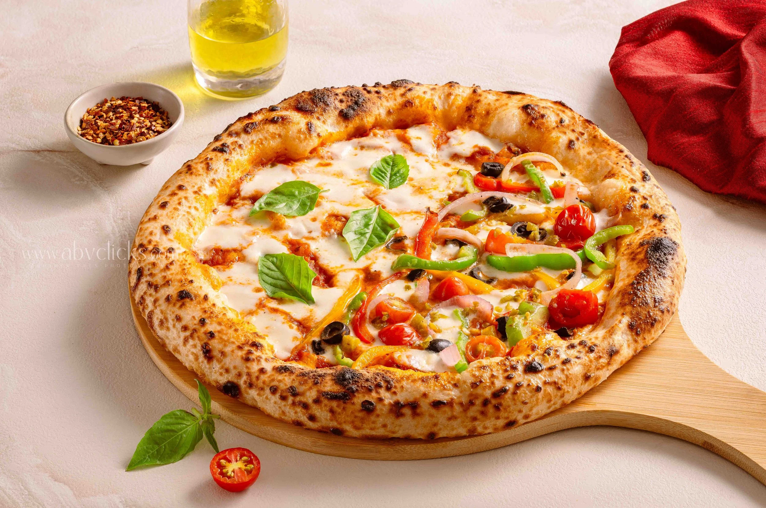 A pizza with a thick crust topped with cheese, basil, cherry tomatoes, black olives, and sliced peppers on a wooden board. A bowl of crushed red pepper flakes, a glass of yellow beverage, and a red cloth are on a light-colored surface.