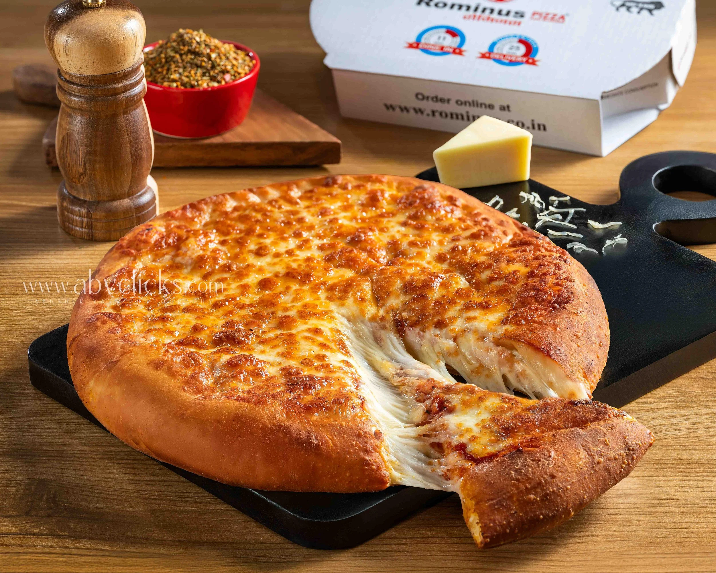 A freshly baked cheese pizza on a black cutting board with a slice being pulled away. In the background, there is a wooden pepper grinder, a bowl of crushed red pepper flakes, and a pizza box with the brand name "Romino's".