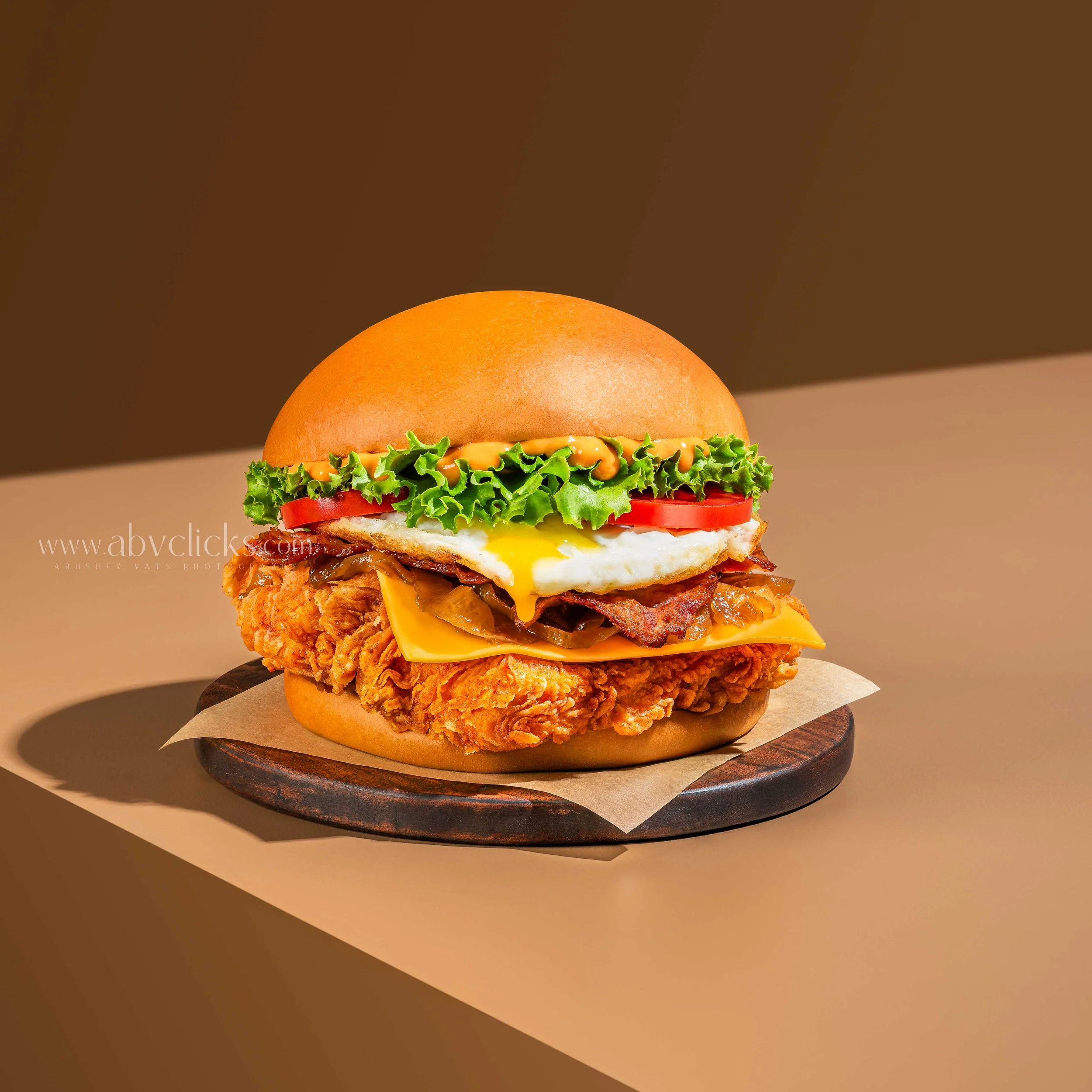 For brand Sandburgs, a close-up of a burger with a crispy fried chicken patty, topped with melted cheese, bacon, lettuce, tomato slices, and a soft bun, placed on a round wooden board against a plain background.