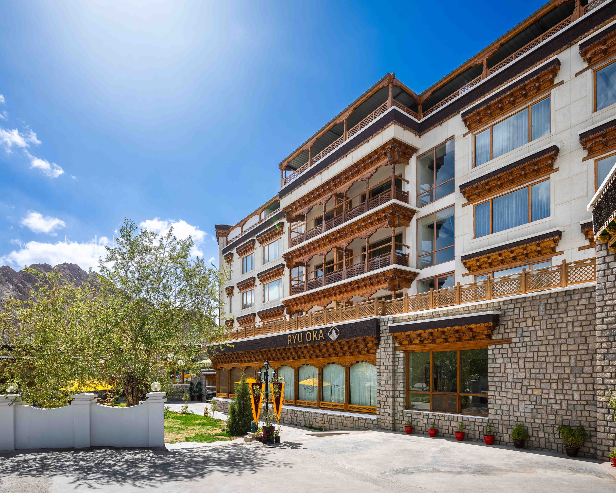 A multi-story building with traditional native architecture style, featuring wooden balconies and carved wooden decorations, with the sign 'RYU OKA' on the exterior, set against a mountainous landscape and a blue sky with some clouds.
