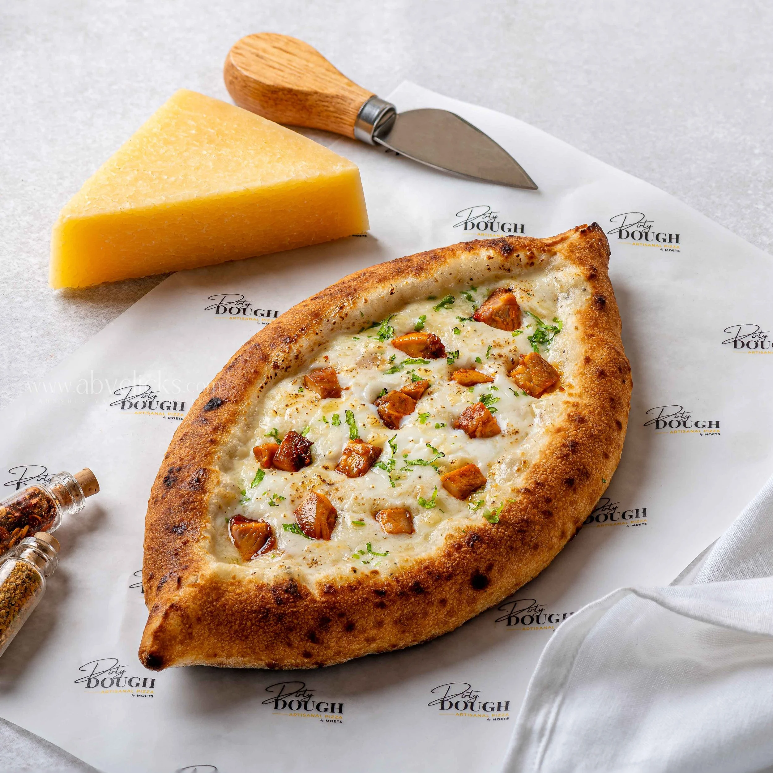 Close-up of a slice of yellow cheese, a pizza with cheese and toppings, and a pizza cutter on branded parchment paper.