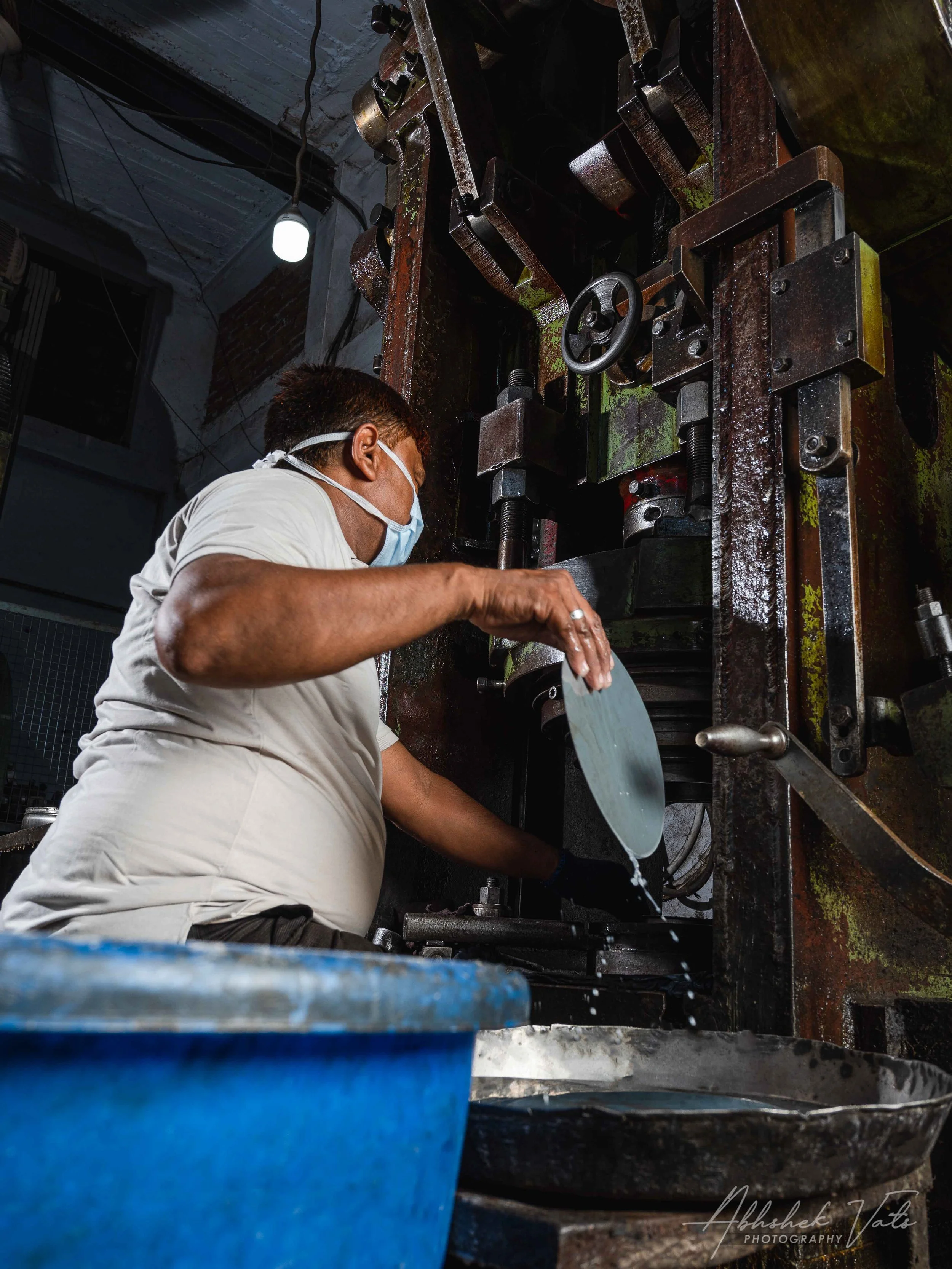 A man wearing a face mask working on a large industrial machine with a wheel and mechanical parts in an industrial setting.