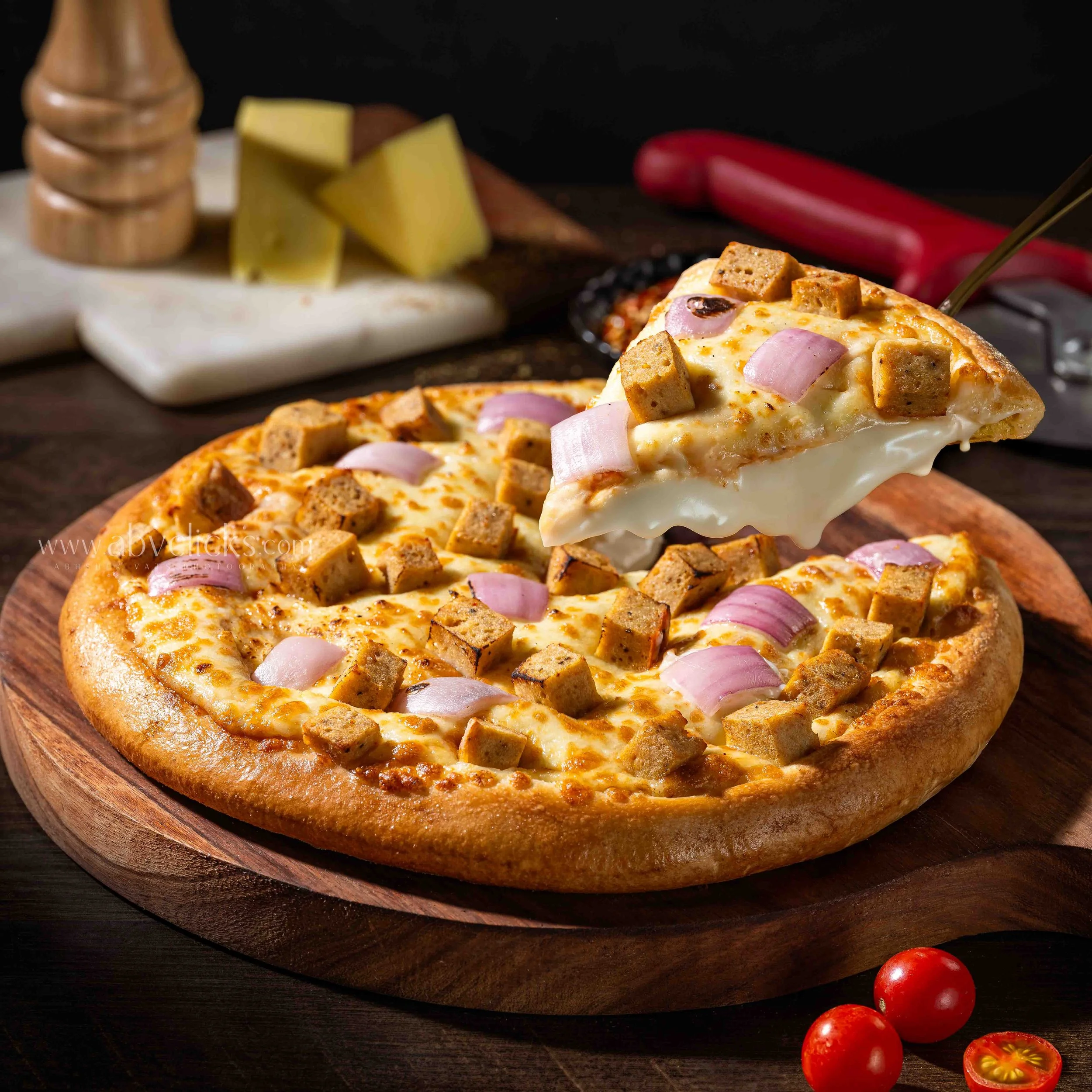 A cheese pizza topped with pepperoni, red onions, and bacon bits on a wooden serving board, with cherry tomatoes nearby.