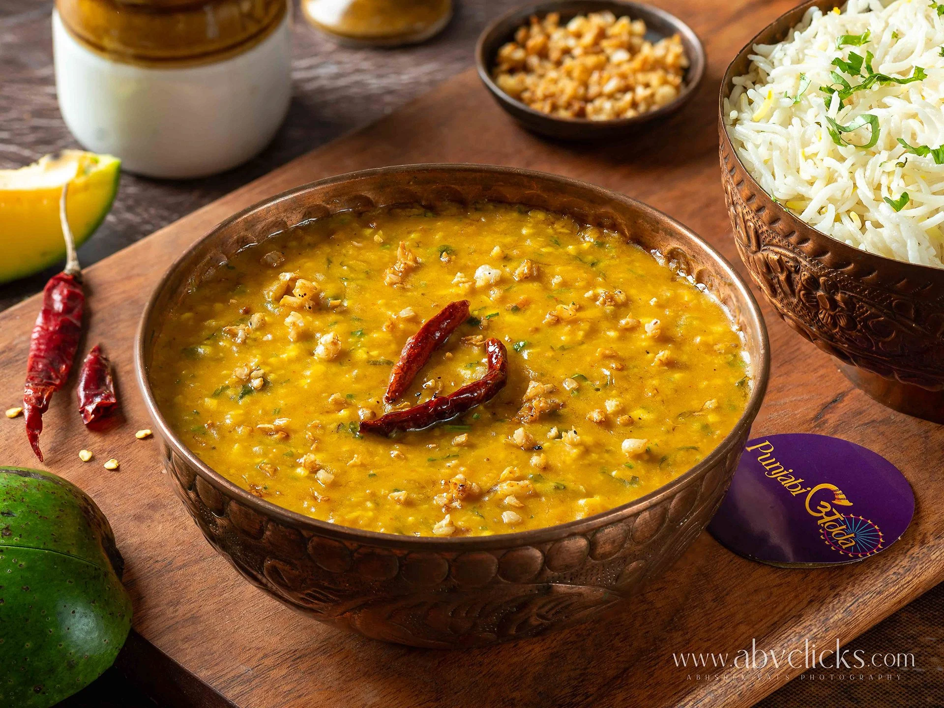 A bowl of Indian yellow lentil dal garnished with dried red chili peppers, with a lemon wedge and green avocado nearby, on a wooden surface.