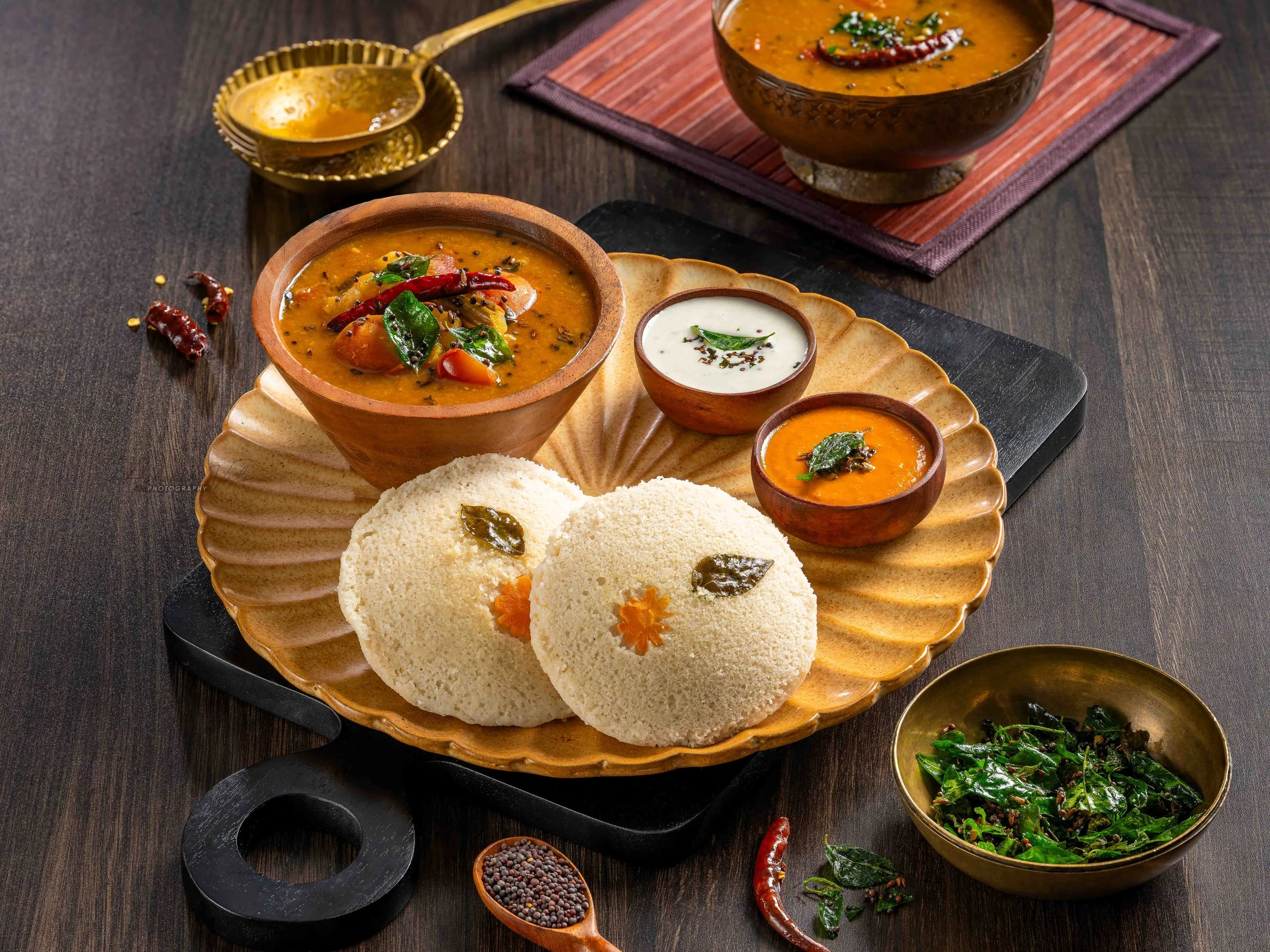South Indian meal with lentil soup, Idli, and various side dishes served on a black platter with small bowls of sambhar.