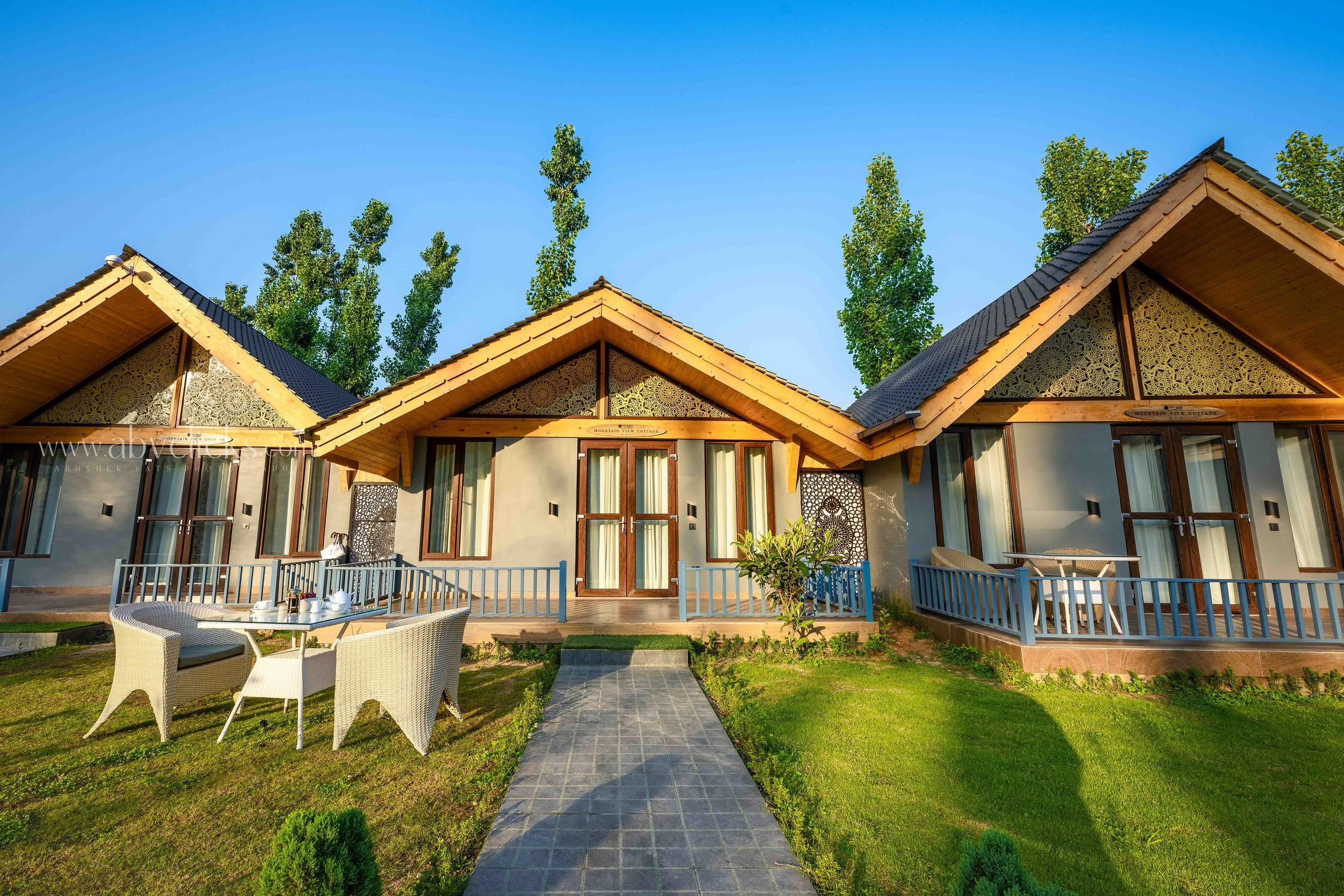 Modern house with a zigzag roof design, surrounded by green grass and trees, featuring a patio with white chairs and outdoor dining set, set against a clear blue sky.