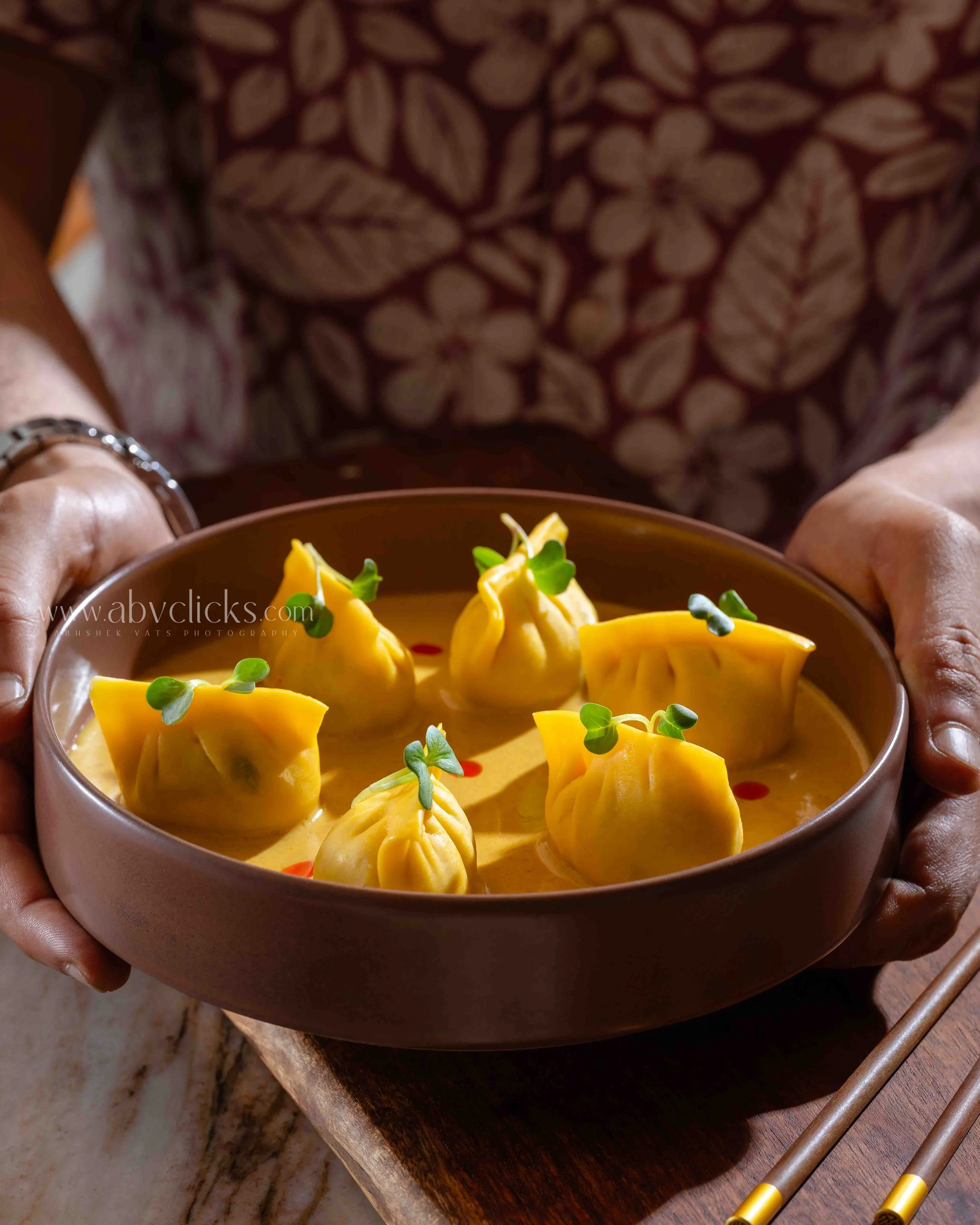 Close-up of hands holding a bowl of yellow dumplings garnished with microgreens, with chopsticks resting on a wooden surface nearby.