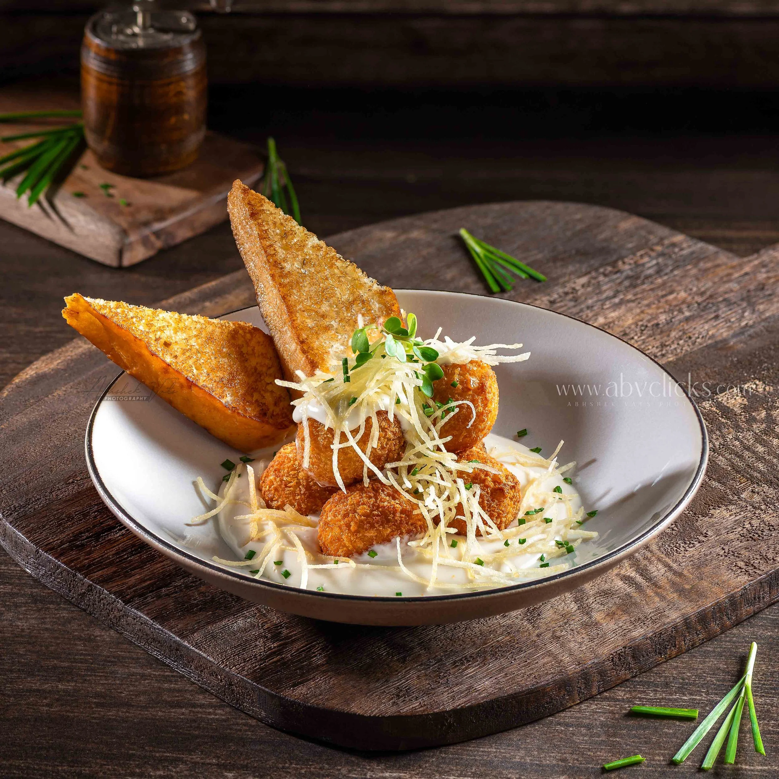 Close-up of a plate with fried cheese sticks topped with shredded cheese and microgreens, on a wooden table with fresh herbs and a dark background.