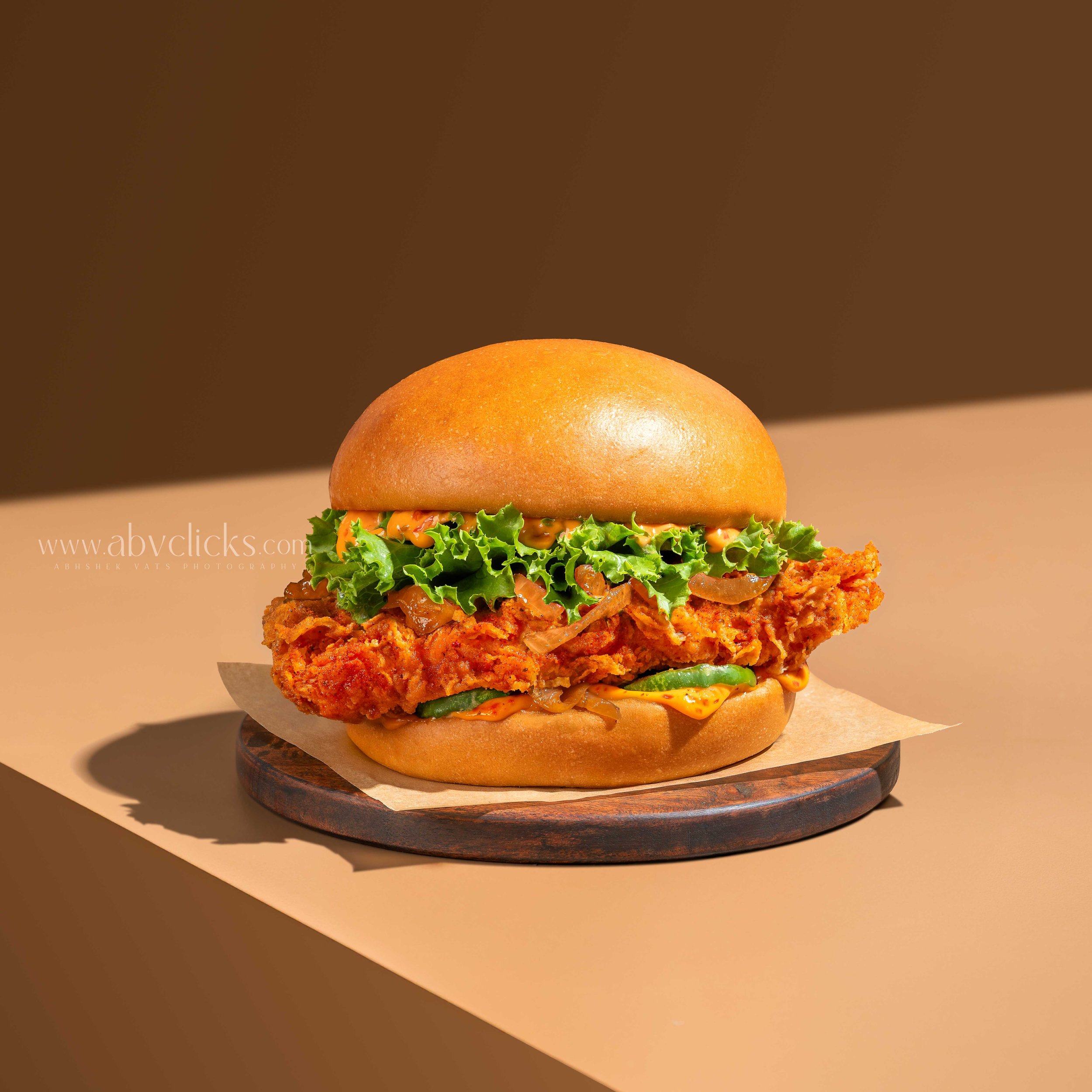 Close-up of a fried chicken sandwich with lettuce, sauce, and a sandwich bun on a wooden board against a brown background.
