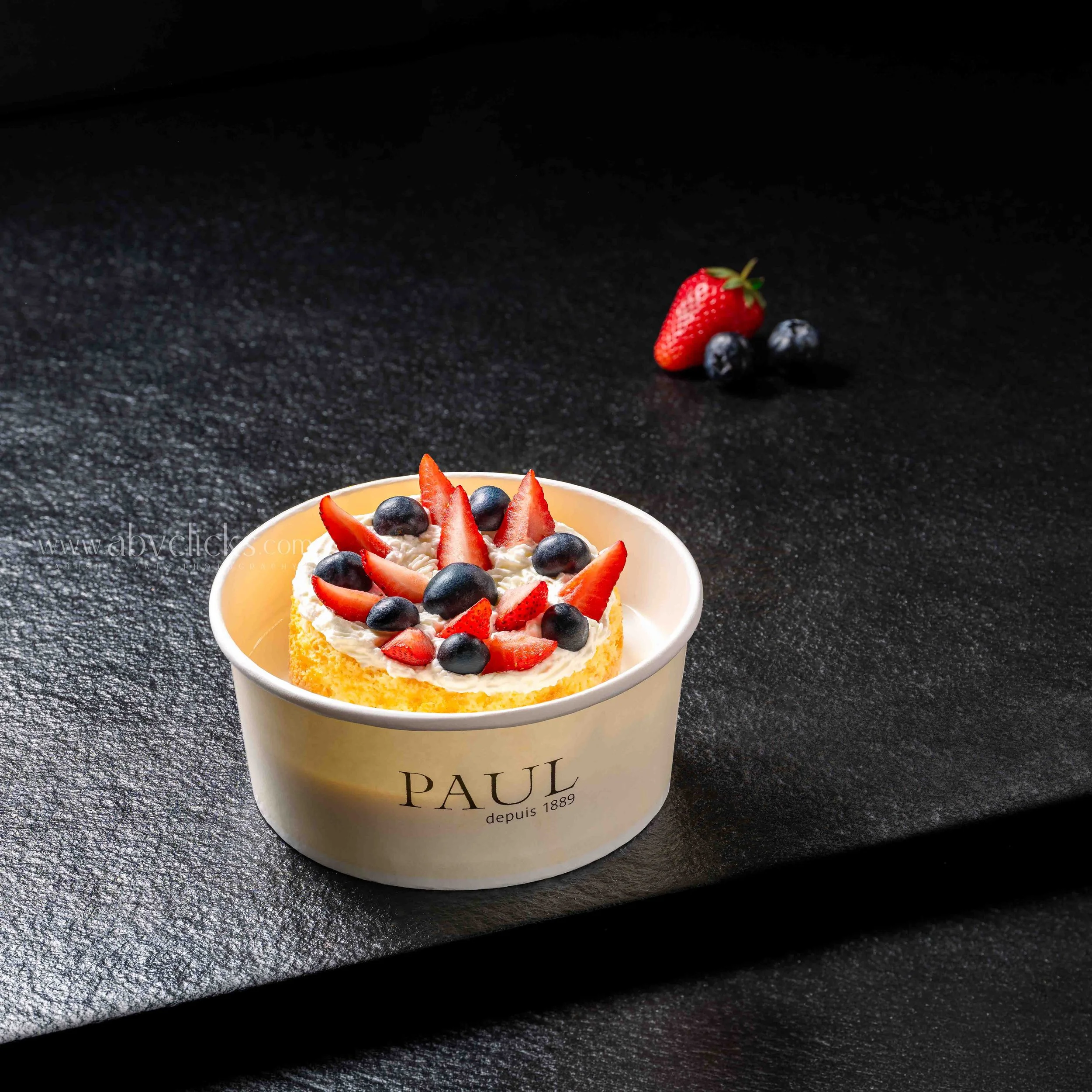 A bowl filled with whipped cream topped with sliced strawberries and blueberries, placed on a dark textured surface. A single strawberry and blueberries are tossed in the air above the bowl.