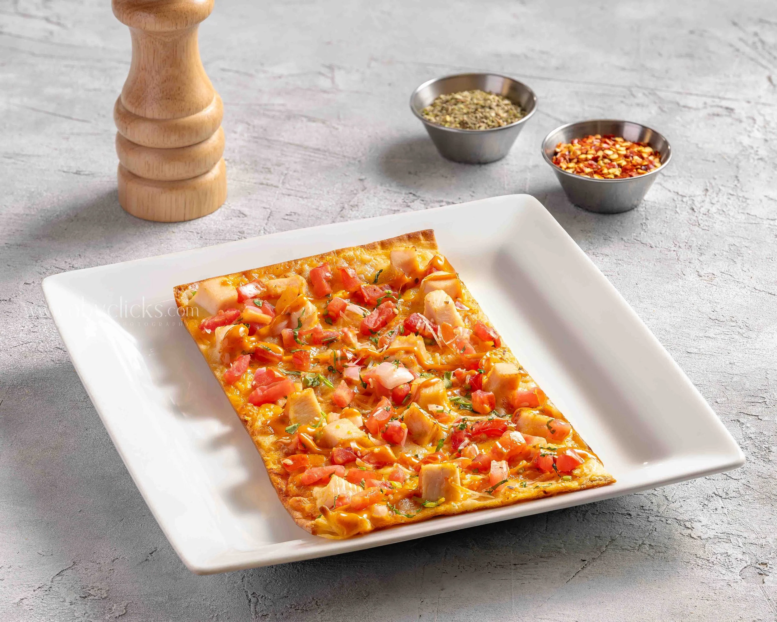 A slice of pizza on a white rectangular plate with toppings including diced tomatoes, chicken, and herbs. In the background, there are small metal bowls containing crushed red pepper flakes and a mixed herbs blend, along with a wooden pepper grinder 