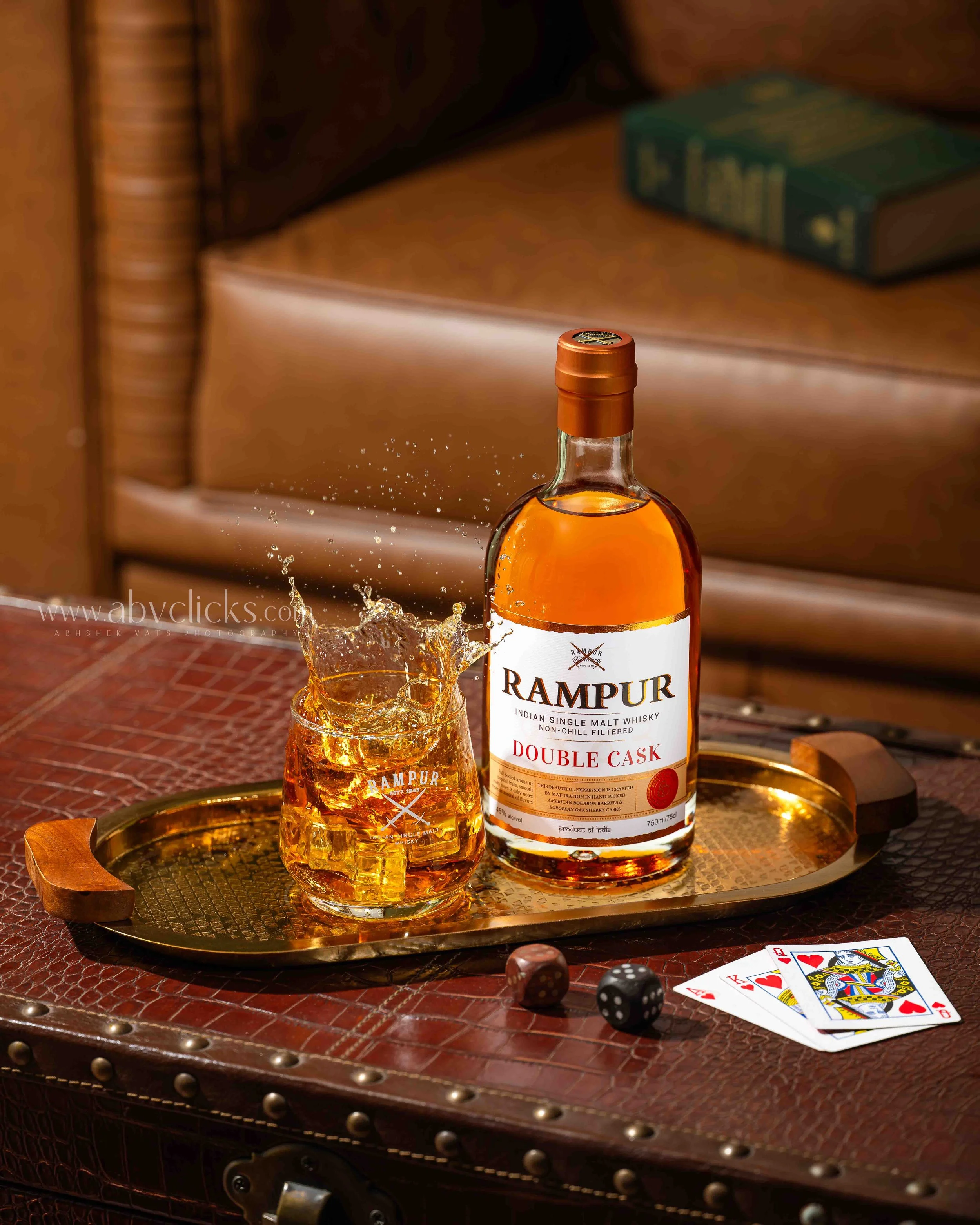 A bottle of Rampur Indian Single Malt Whisky with a glass of whisky beside it, placed on a round brass tray on a wooden table, with playing cards and dice nearby, and a leather armchair with books in the background.