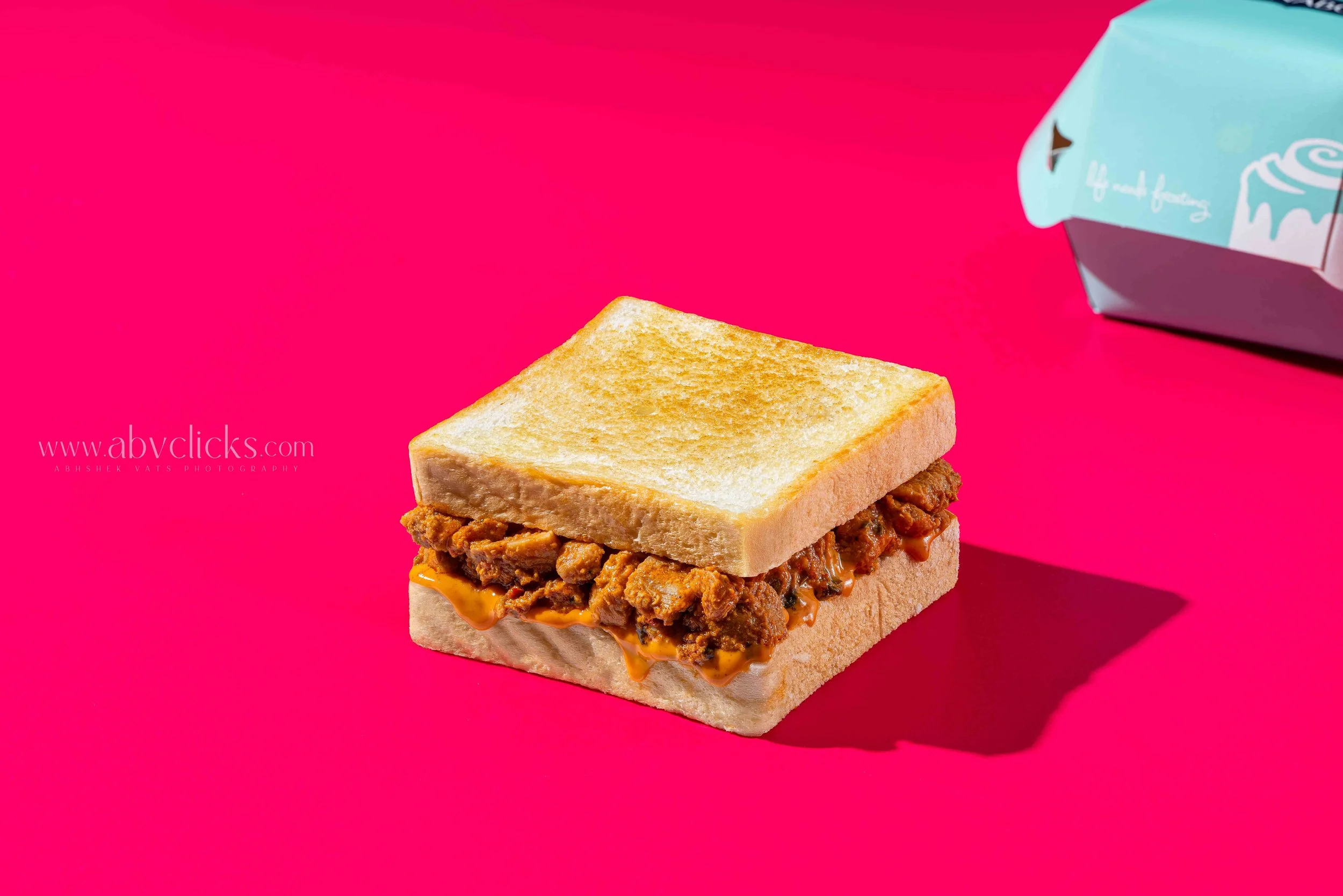 A close-up of a sandwich with chopped meat and melted cheese between two slices of white bread, on a bright pink background, with a blue and white packaged snack in the corner.