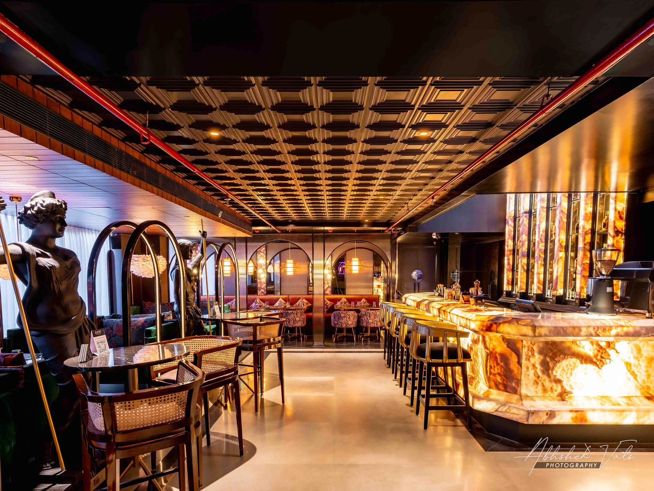 Interior of a stylish bar or lounge with statues, a marble bar counter with barstools, warm lighting, and decorative walls with paintings and mirrors.