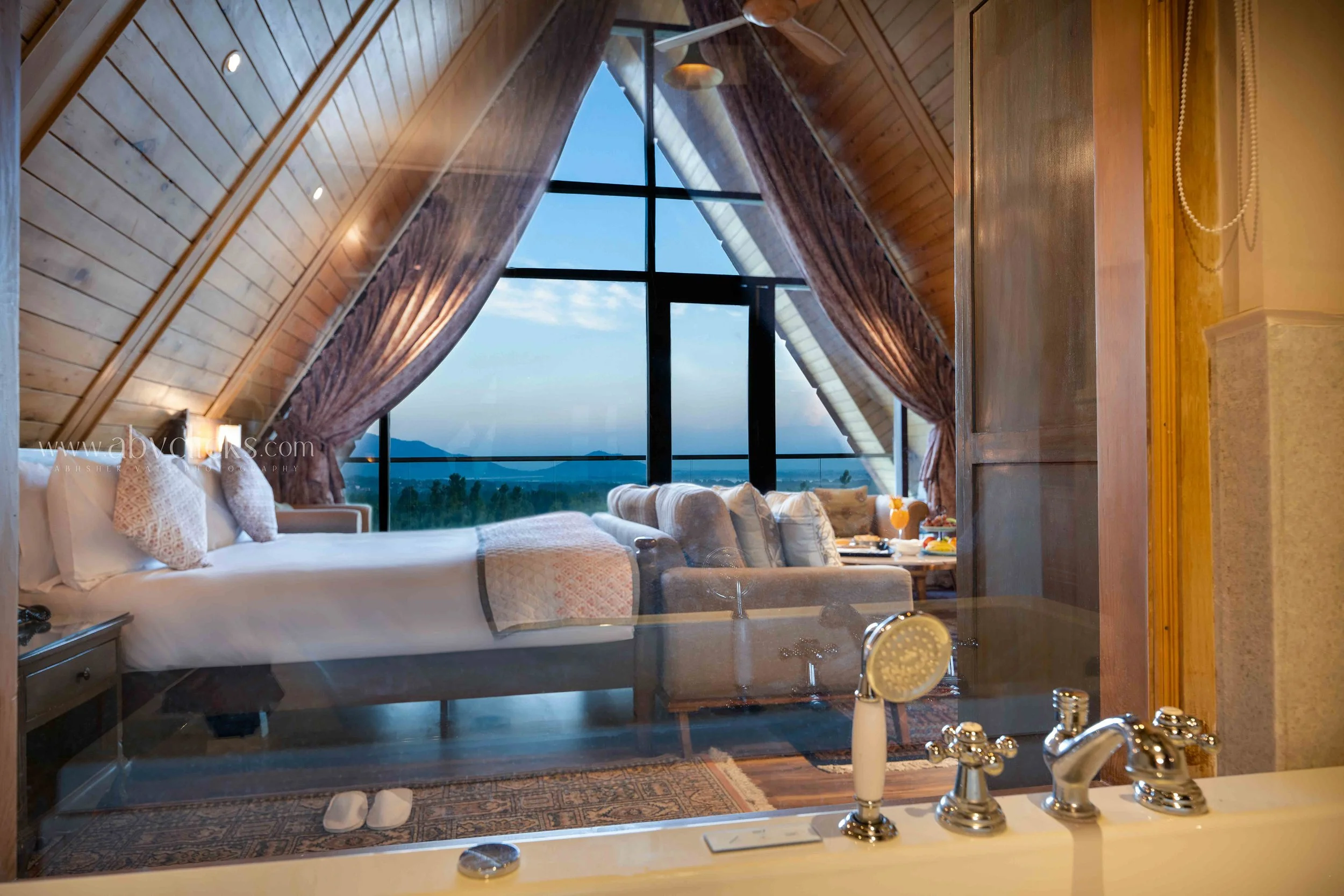 A bedroom with a large window, rustic wood ceiling, and a bed with white and patterned pillows. There are curtains hanging on each side of the window, providing a view of a landscape with mountains and trees outside. The room has warm lighting and co
