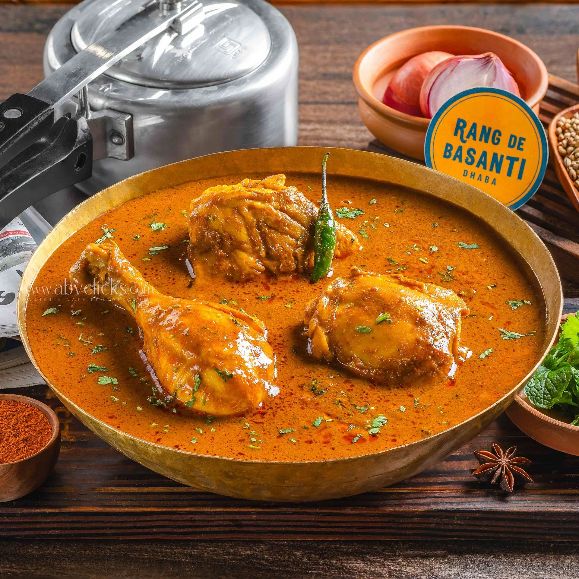 A bowl of Indian chicken curry garnished with herbs and a green chili, with side dishes of spices, onions, and coriander on a wooden table.