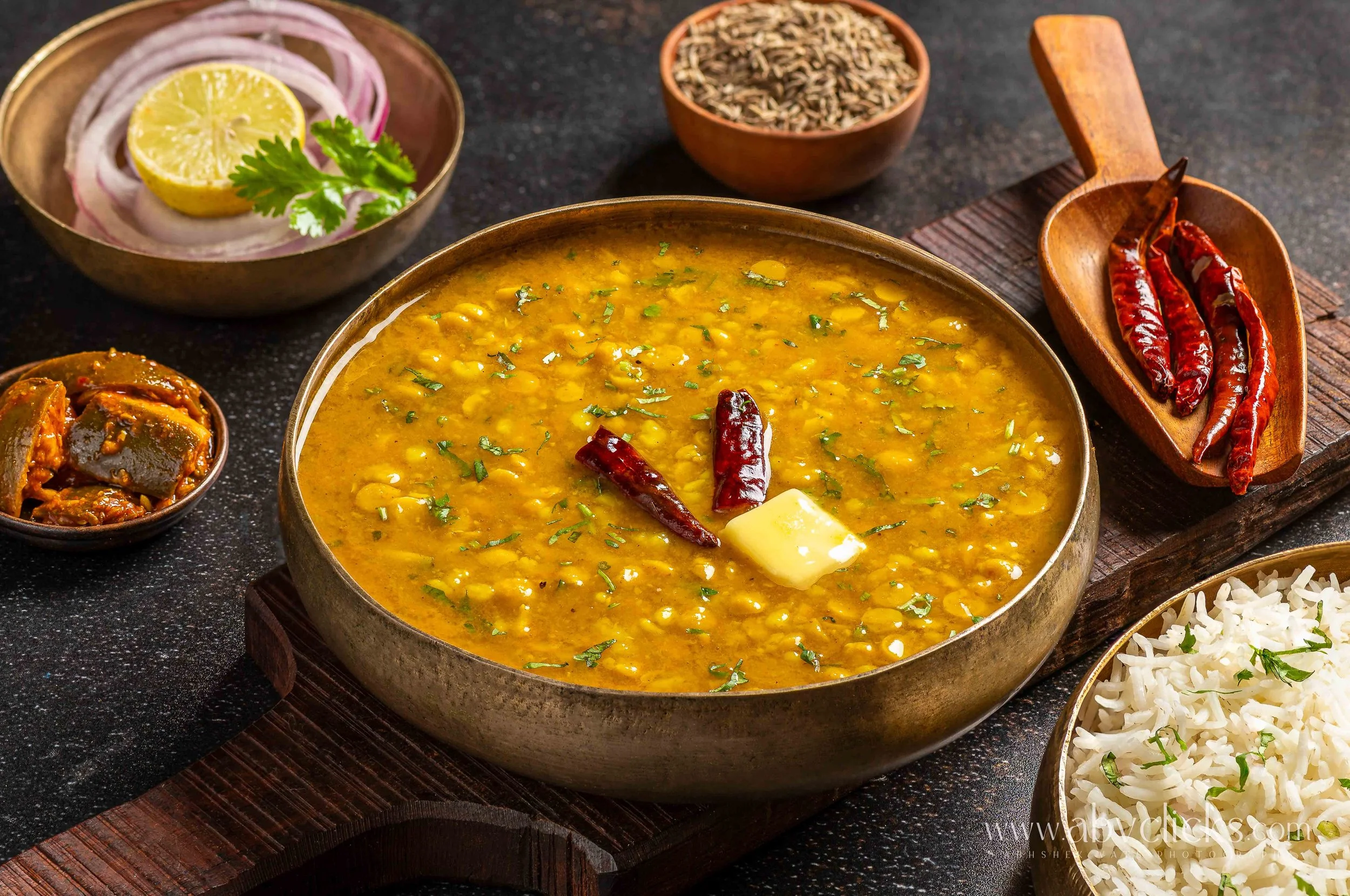 A bowl of yellow lentil curry garnished with cilantro, dried red chili peppers, a small dish of pickles, a bowl of chopped red onion and lemon, a small bowl of cumin seeds, and a bowl of cooked rice on a dark surface.