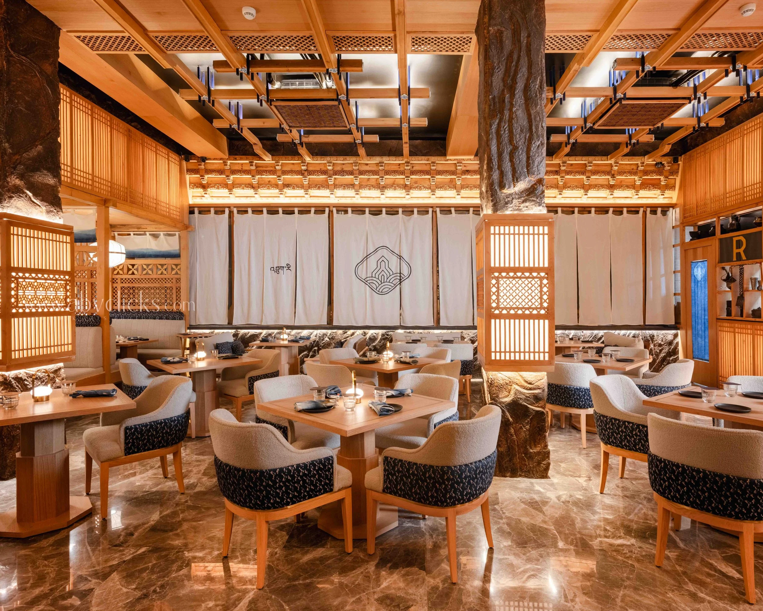 Interior of a restaurant with wooden decor, round tables set with glasses, napkins, and plates, and plush chairs with patterned cushions. Large stone columns are visible, and the ceiling has wooden beams and a decorative lattice structure.