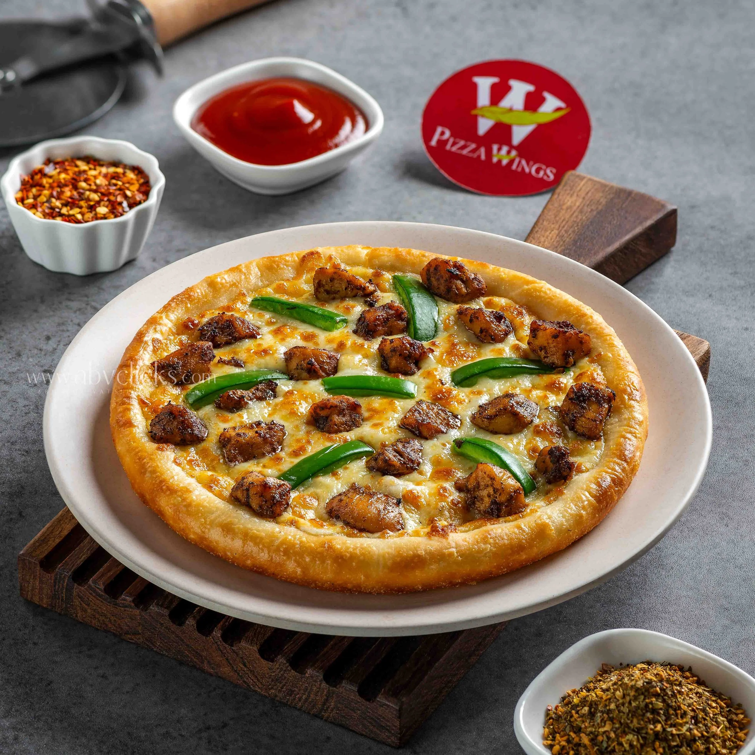 Close-up of a pepperoni pizza with green peppers on a white plate, accompanied by bowls of crushed red pepper flakes, tomato sauce, and a dry herb mixture, on a dark surface.
